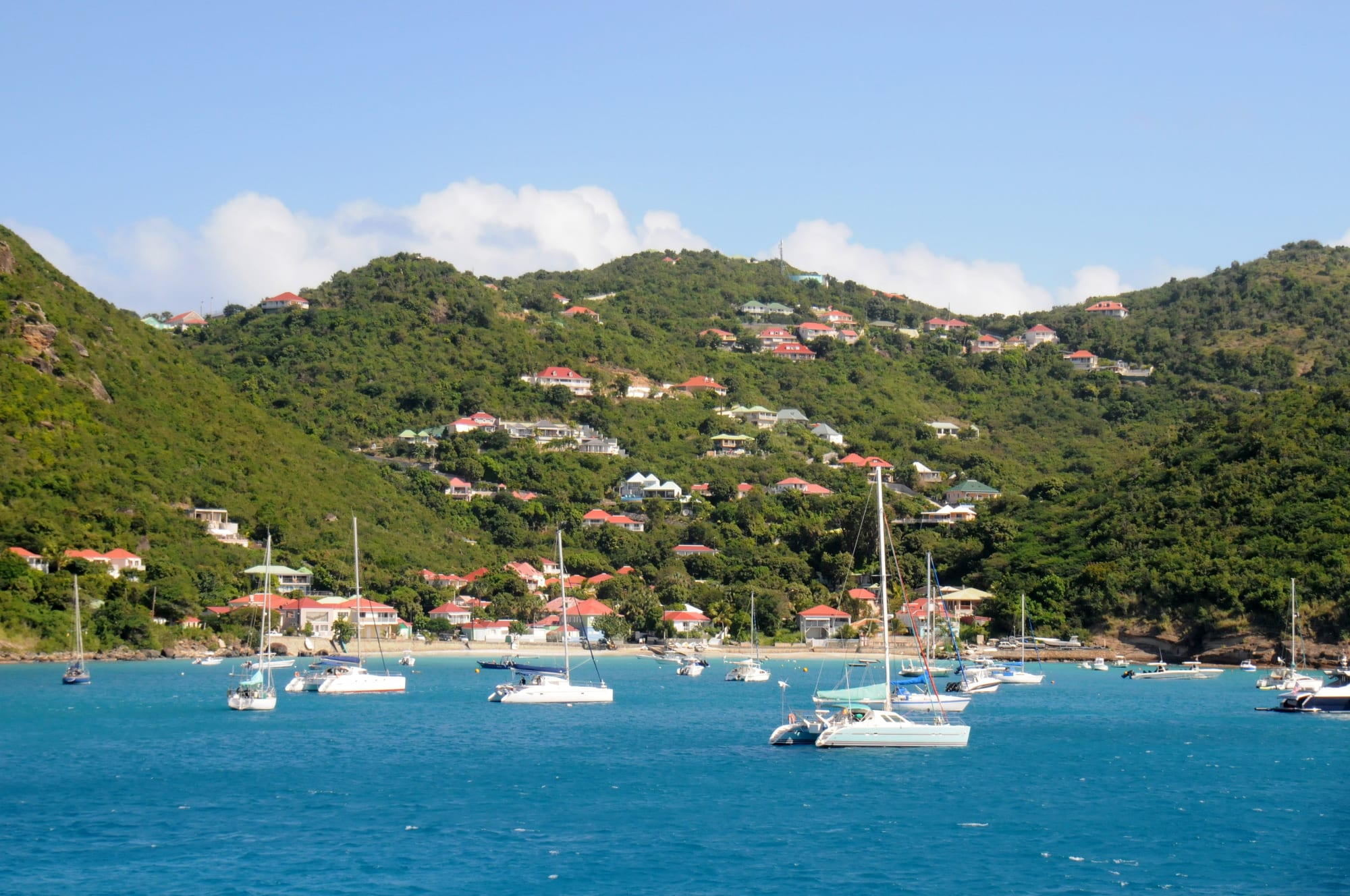 A cluster of white and pastel-roofed houses along a small bay, with sailboats moored in the water and green hills rising behind