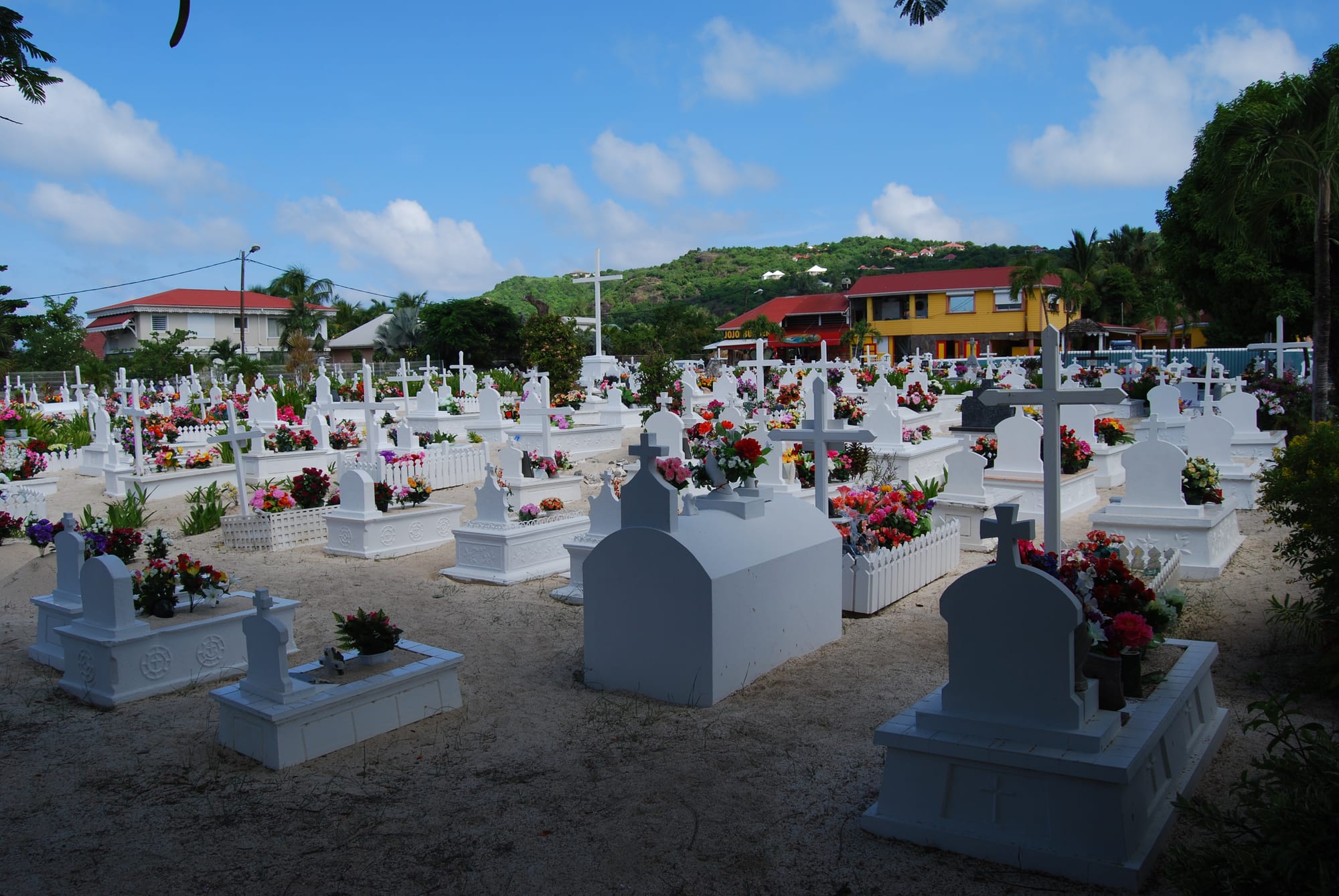 A sandy cemetery crowded with white-painted tombs, crosses and small picket fences, almost every grave decorated with bright pink, red and yellow flowers, with houses and green hills behind