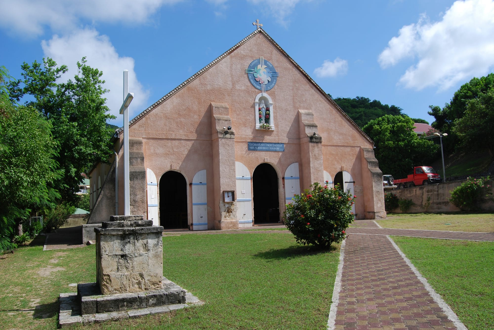 A simple pink-plastered church with a pointed gable, three arched doorways with pale blue shutters, a statue in a niche and a round emblem above, fronted by a lawn, a stone pedestal and a tall white cross