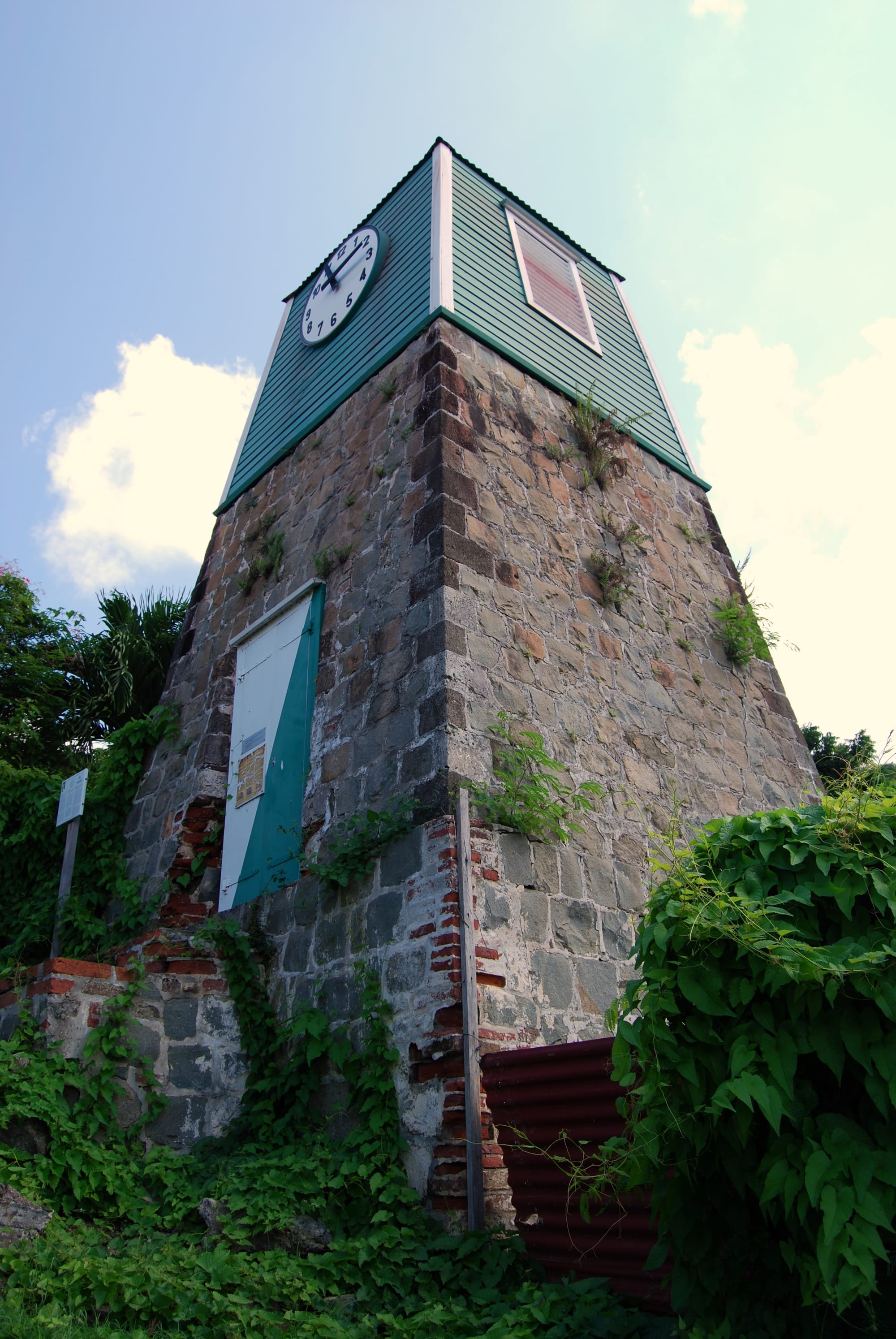 A square tower of rough grey and brown stone with a painted door at its base, topped by a green weatherboarded upper storey with a large white clock face, surrounded by climbing plants