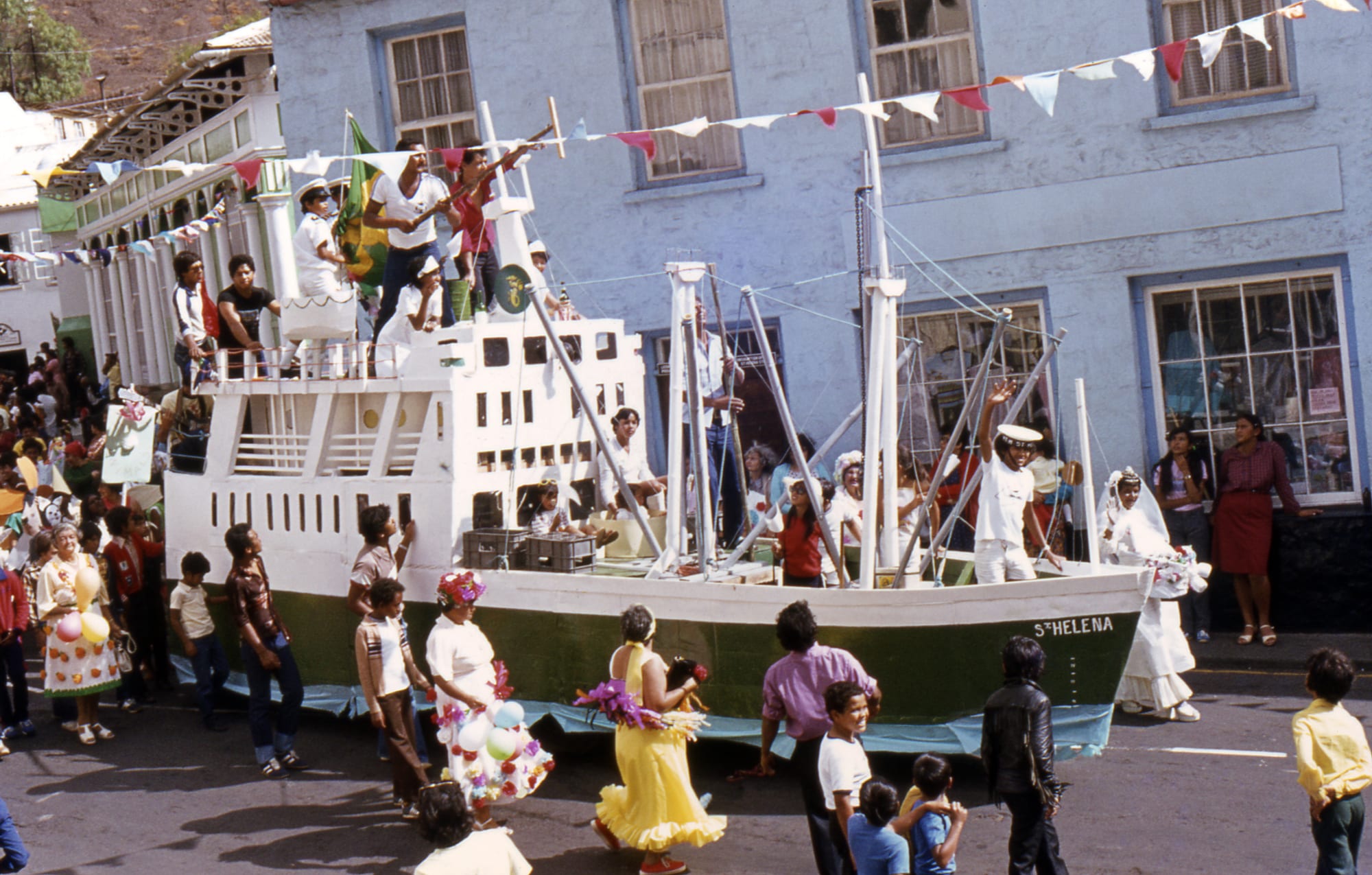 A parade float built as a white and green ship with masts and bunting, crewed by people in white, moving along a street past crowds in costume and onlookers