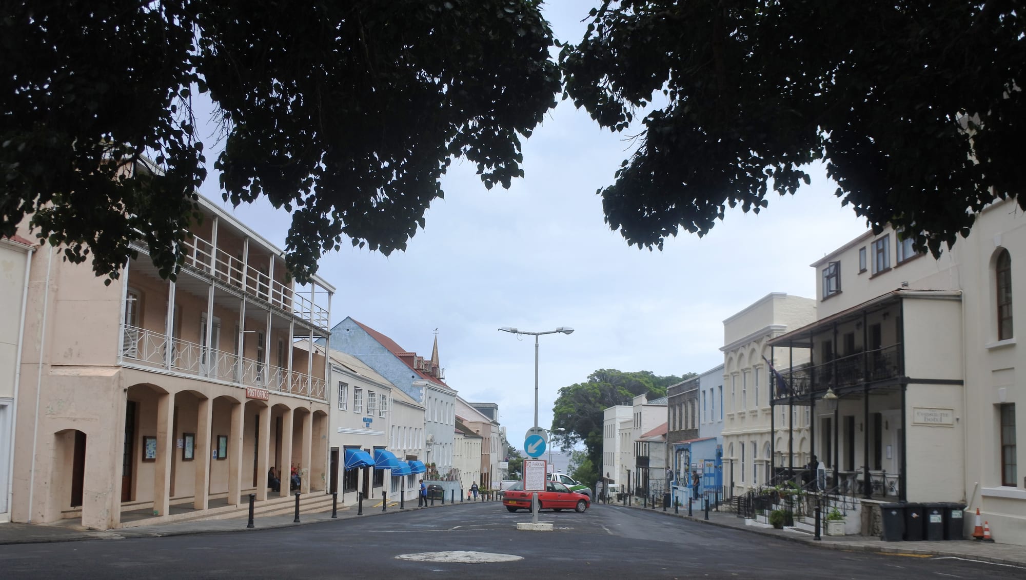 A wide street lined with pale Georgian buildings with balconies and arcades, framed by dark overhanging trees, with a red car in the middle distance