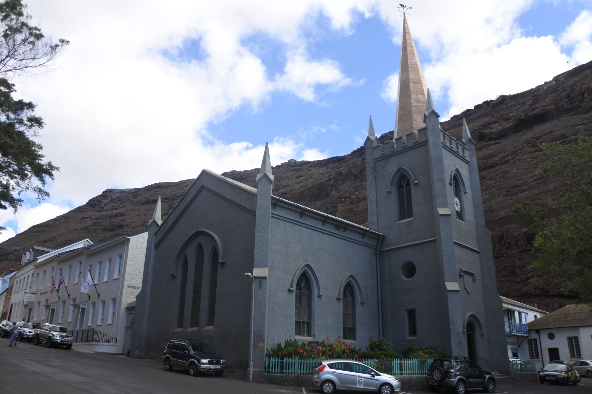 A grey stone church with pointed windows and a tall square tower topped by a spire, with cars parked along the street in front and a bare brown cliff rising behind
