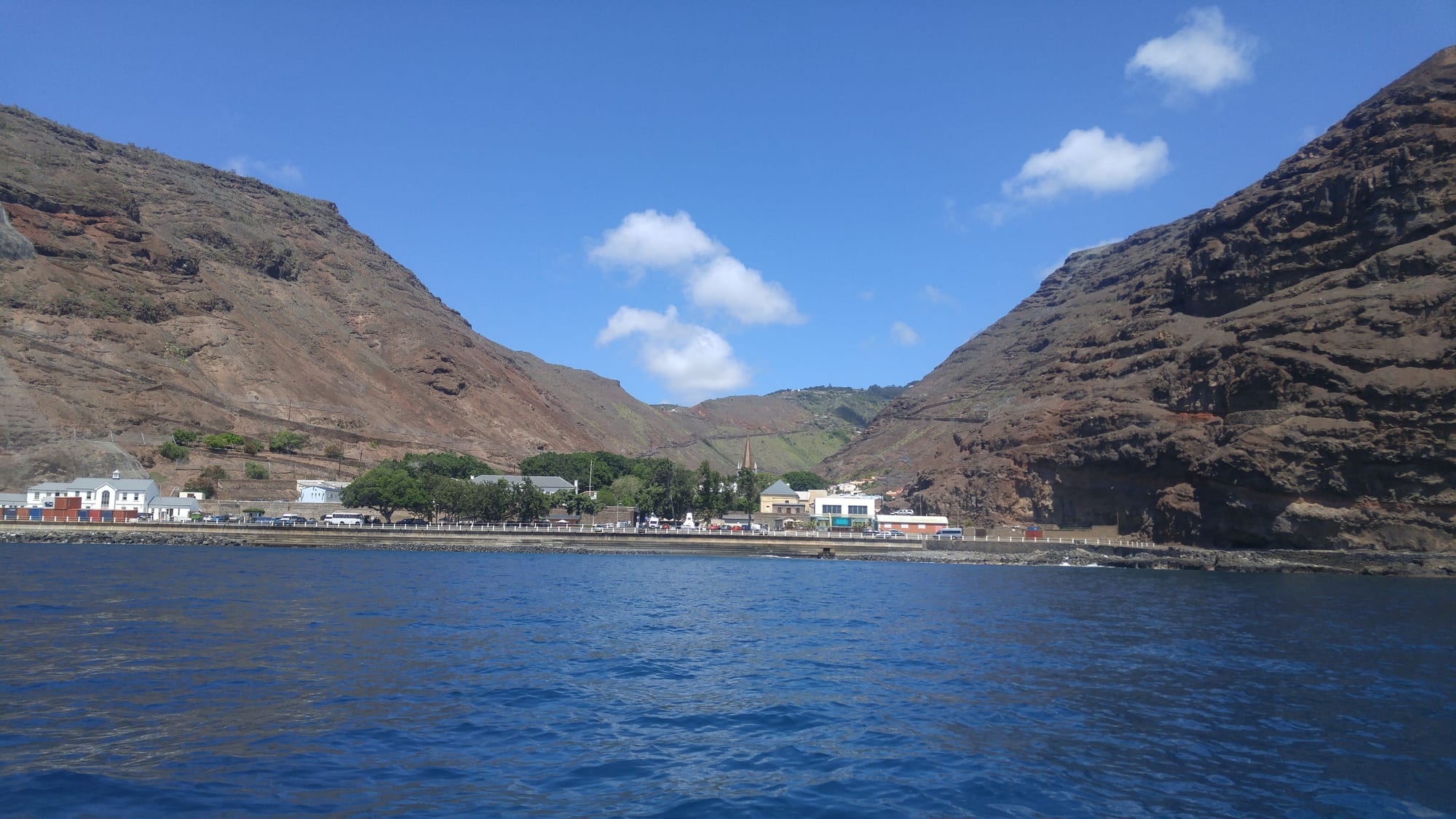 A small town of pale buildings squeezed into the mouth of a steep, bare valley opening onto the sea, seen from a boat offshore