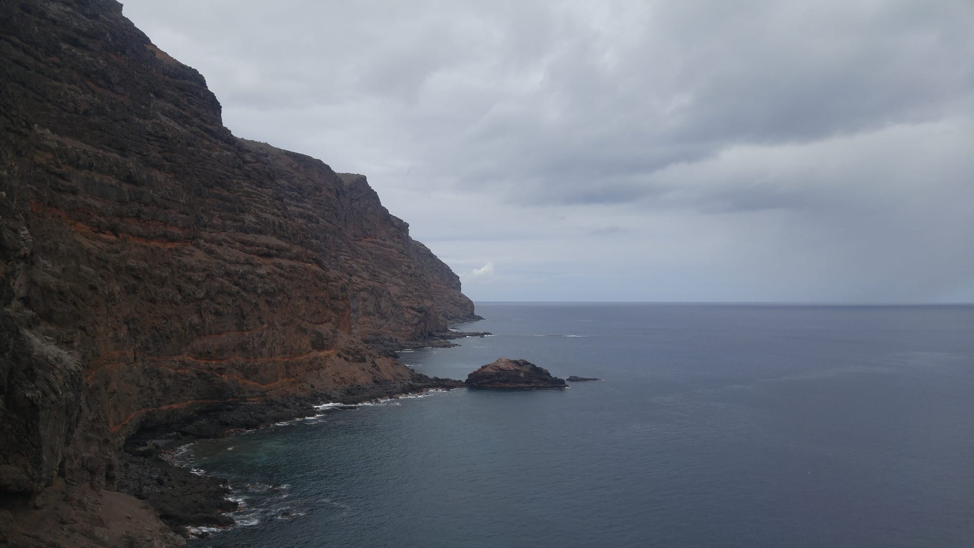 A rugged volcanic coastline with dark red-streaked cliffs and a small offshore rock