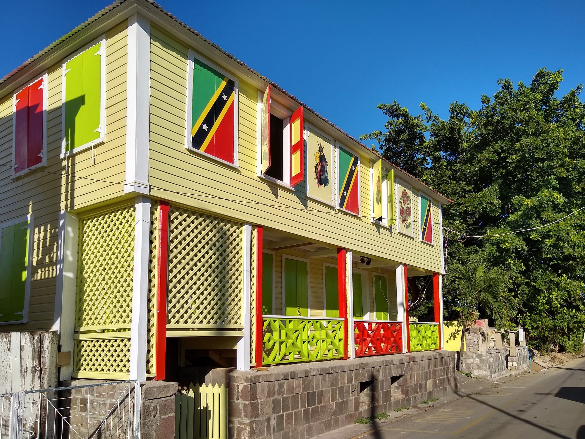 A two-storey wooden gingerbread-style house painted yellow with green and red shutters and a covered porch