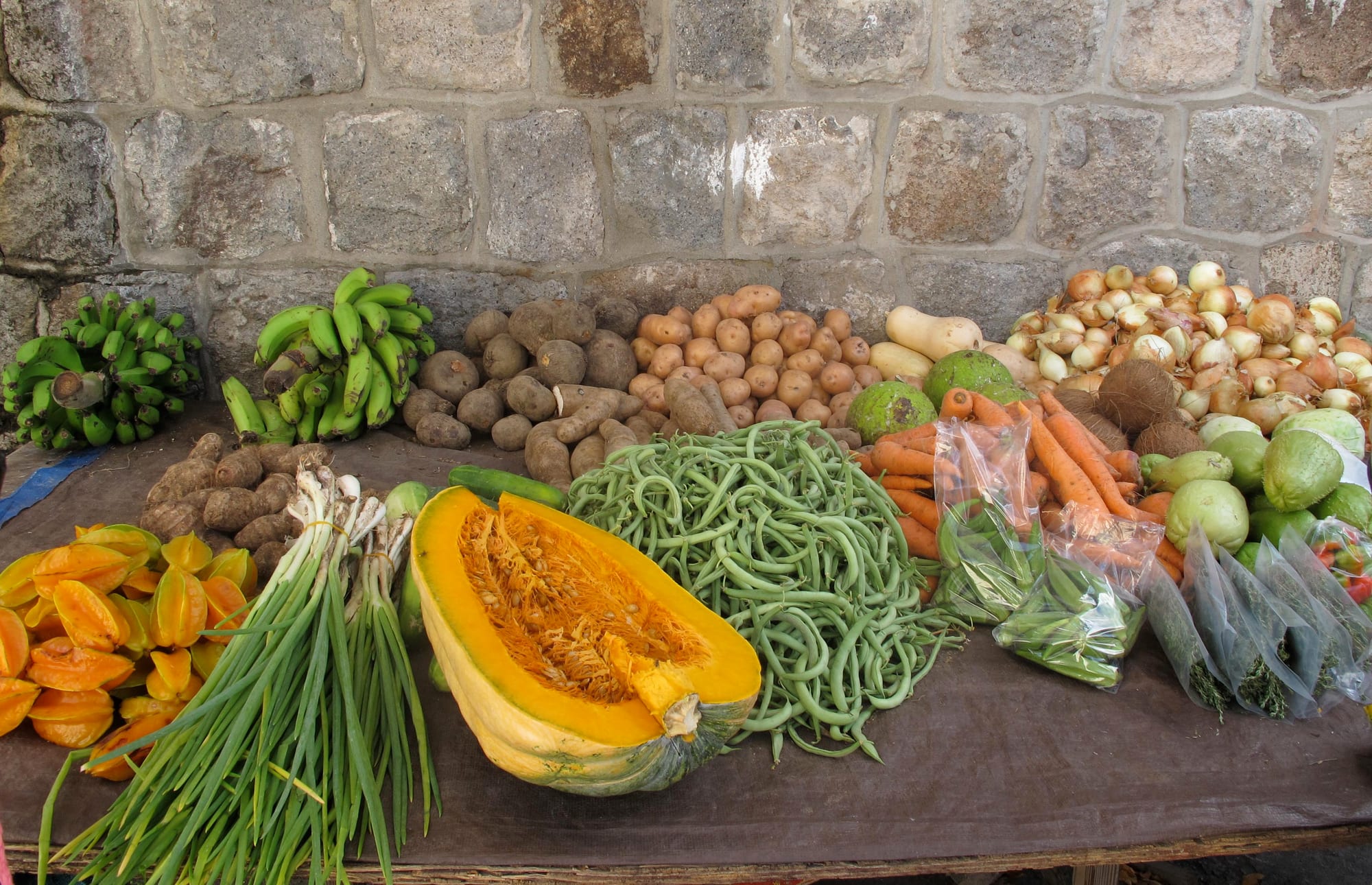 A market stall table piled with bananas, dasheen, pumpkin, carrots and onions against a stone wall