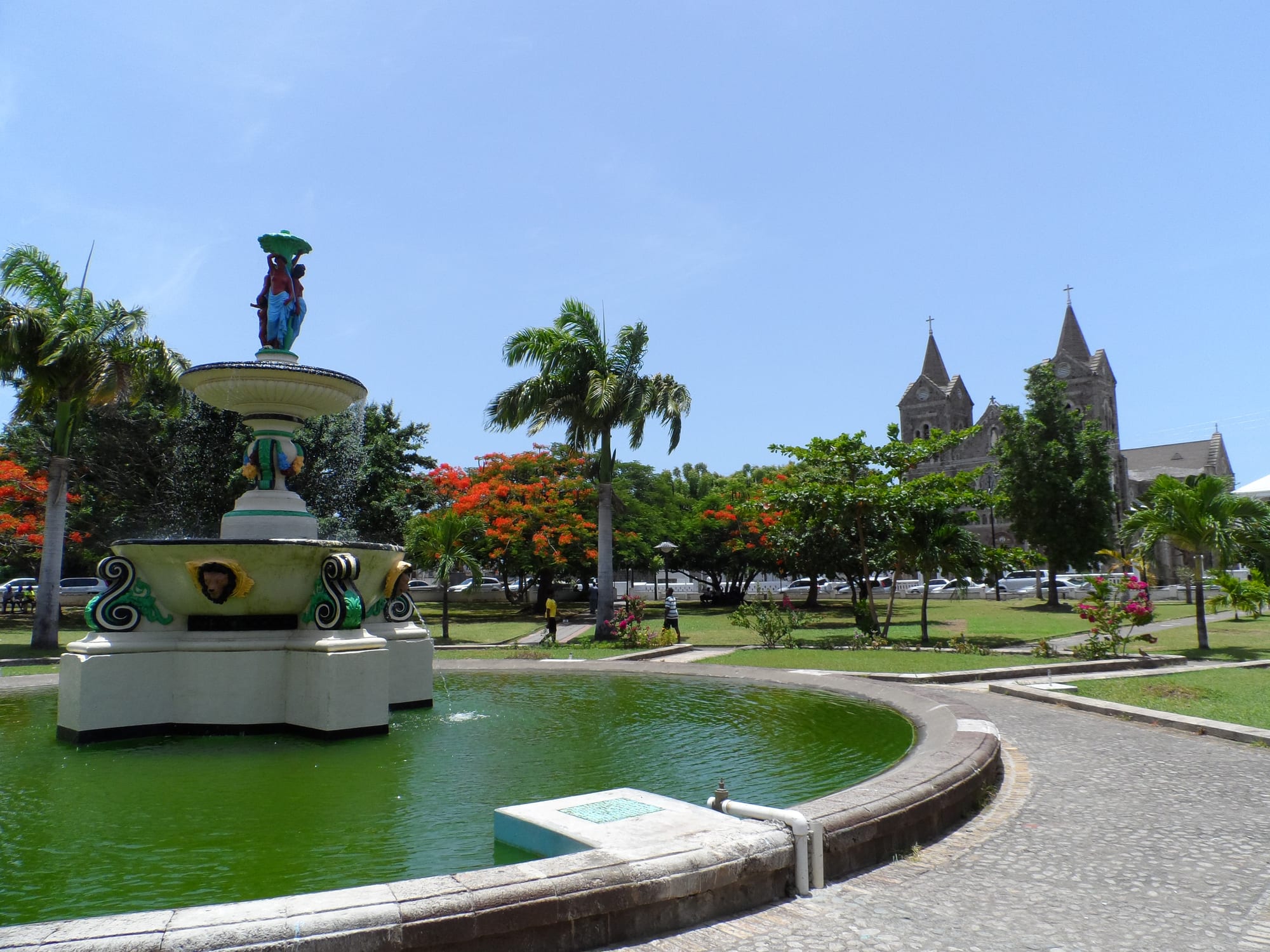 An ornate fountain in a round pool in a landscaped square with palms and flame trees, with the twin spires of a stone cathedral visible beyond the trees