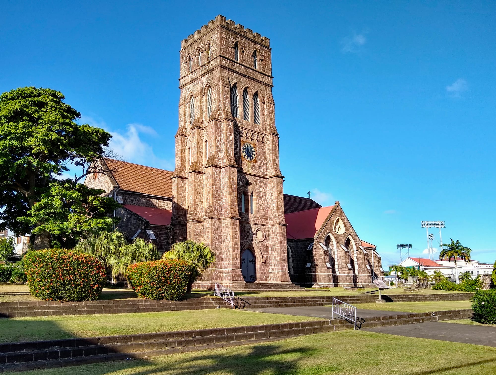 A large church of reddish-brown cut stone with a tall square clock tower and red roofs, set behind clipped flowering hedges and lawns under a clear blue sky