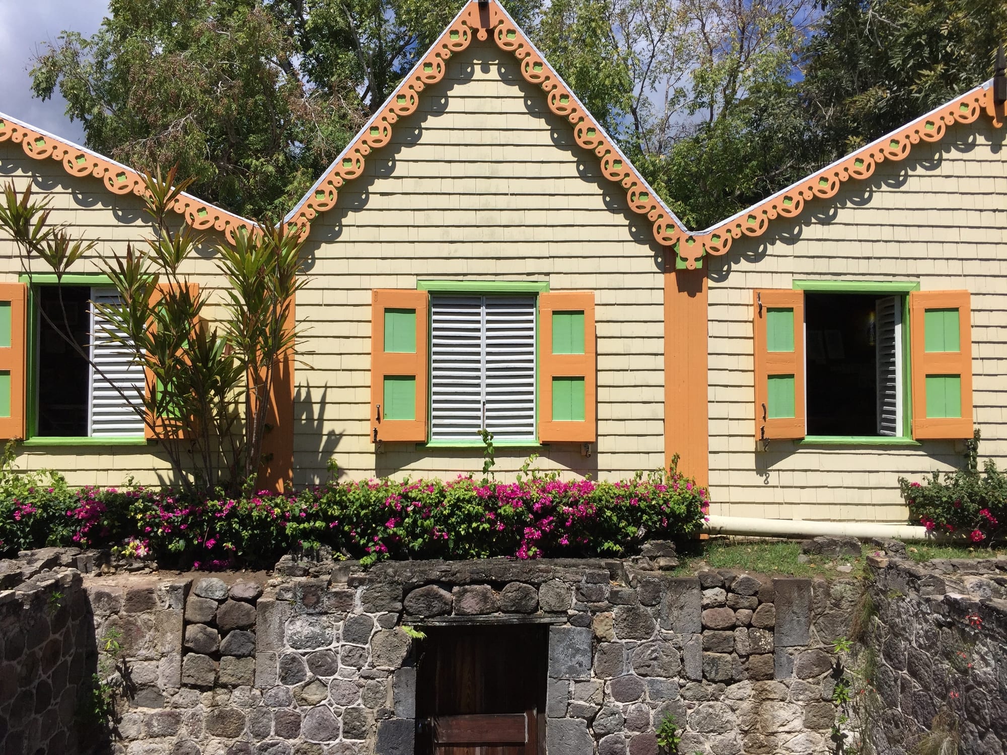 A pale shingled wooden building with orange carved gingerbread trim along its gables and orange and green shutters, above a low stone wall and a bed of pink bougainvillea