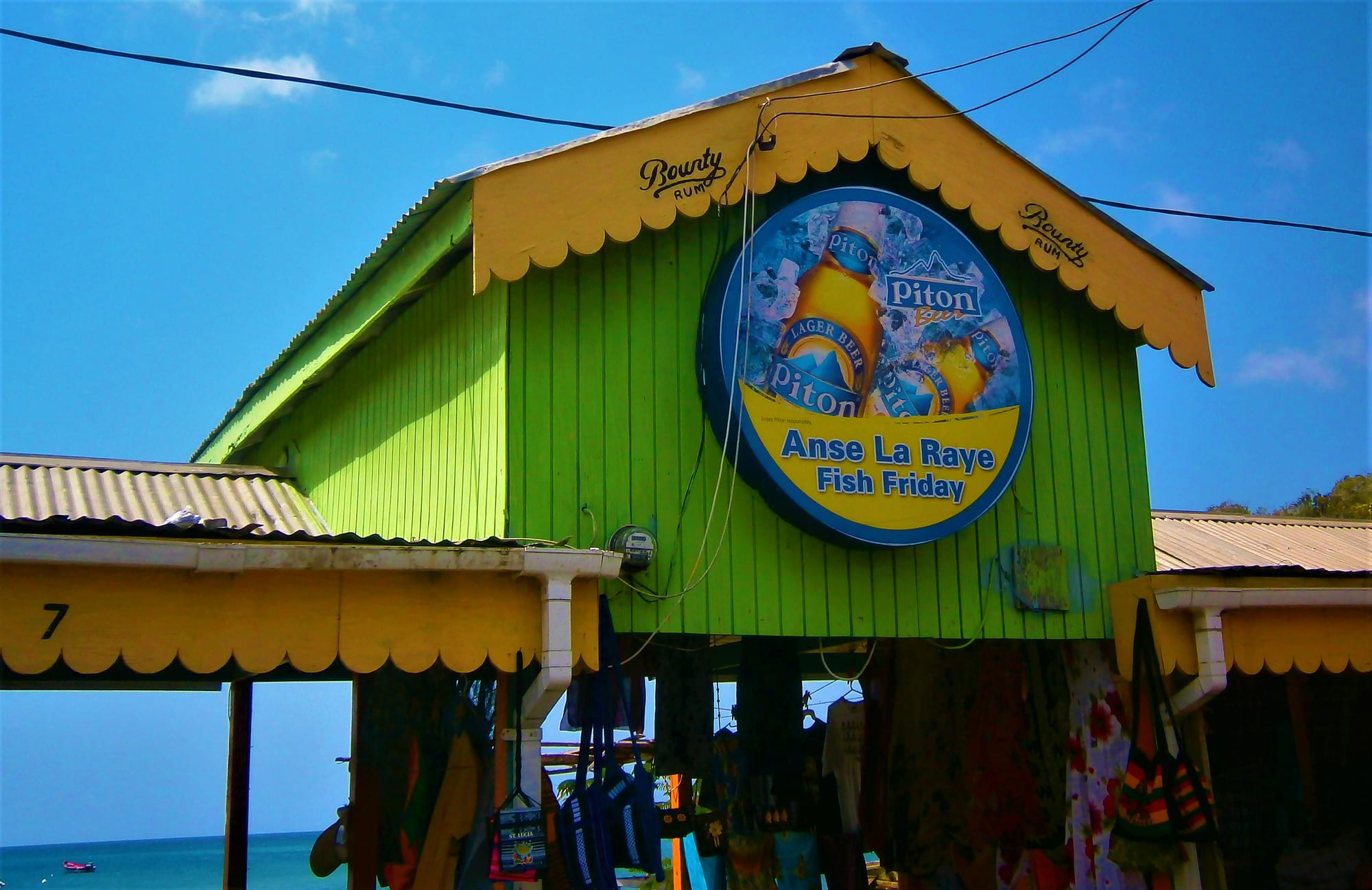 A bright green and yellow wooden stall building with scalloped eaves and a large round sign reading Anse La Raye Fish Friday, with bags and clothing hung for sale beneath it and the sea behind