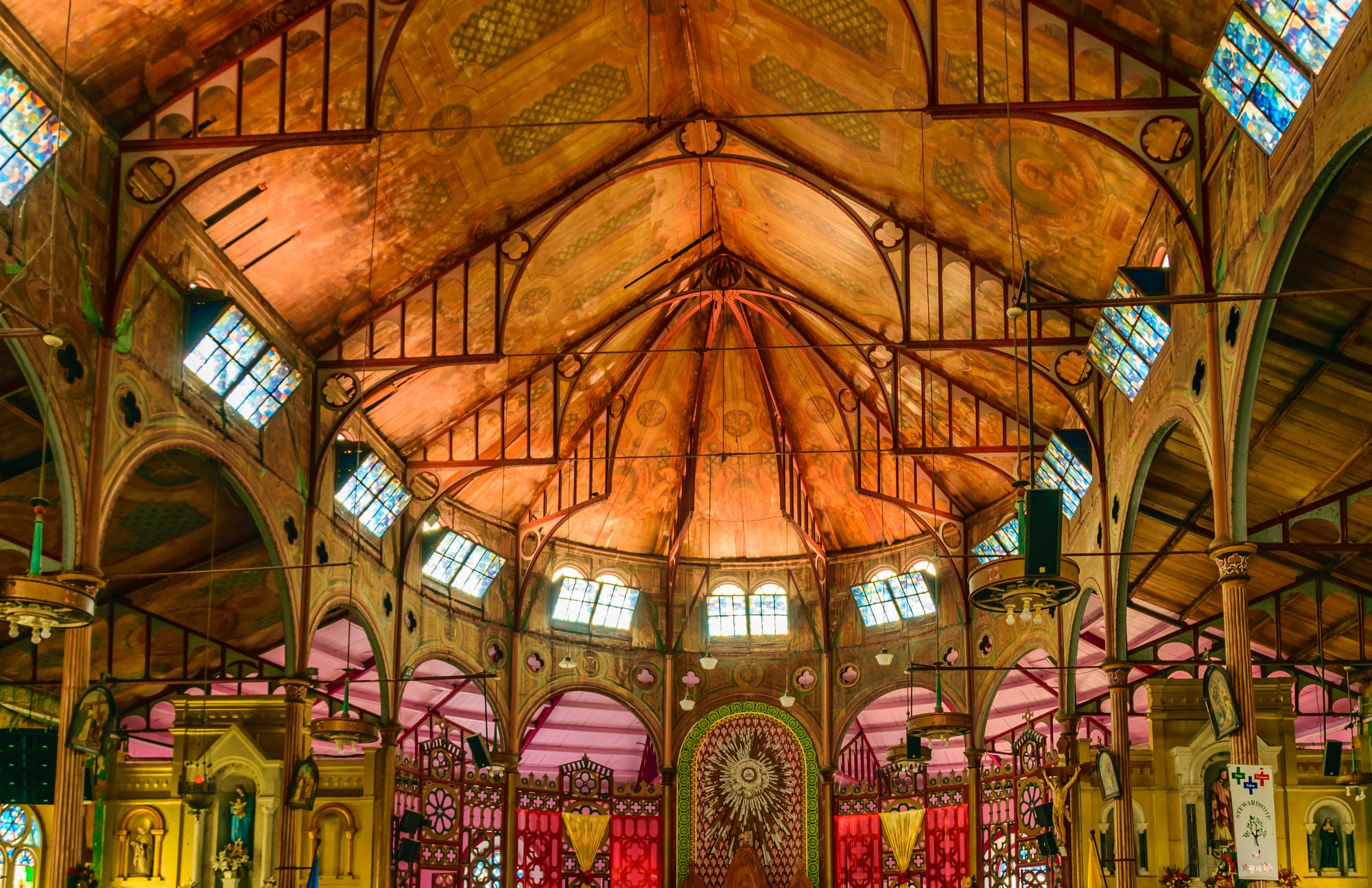 The interior of a large church with a vaulted wooden ceiling painted in warm orange and gold, rows of small coloured windows and a brightly painted red, pink and yellow sanctuary