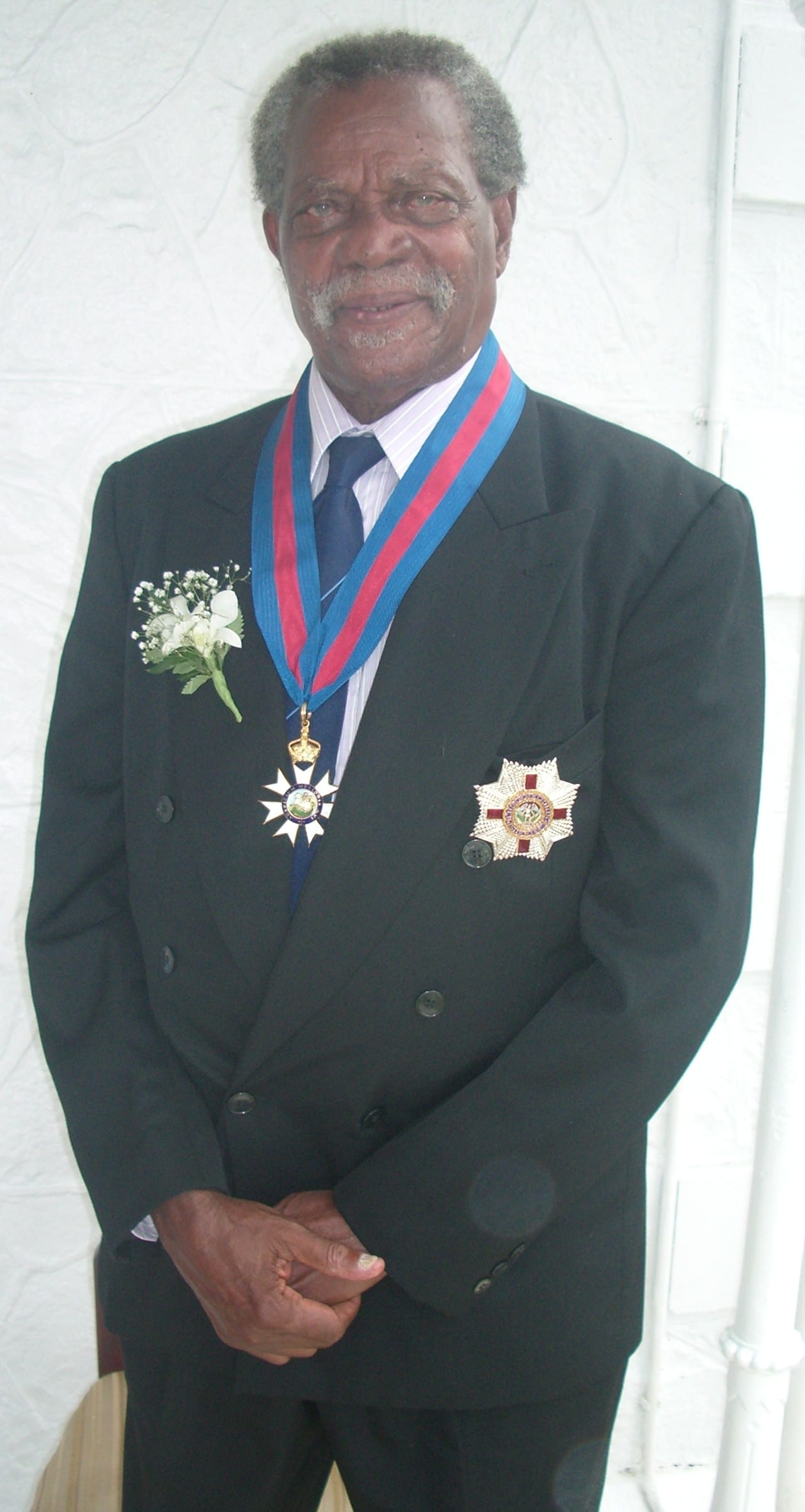 An elderly man with grey hair and a moustache in a dark double-breasted suit, wearing a medal on a blue and red ribbon around his neck, a silver breast star and a white flower buttonhole