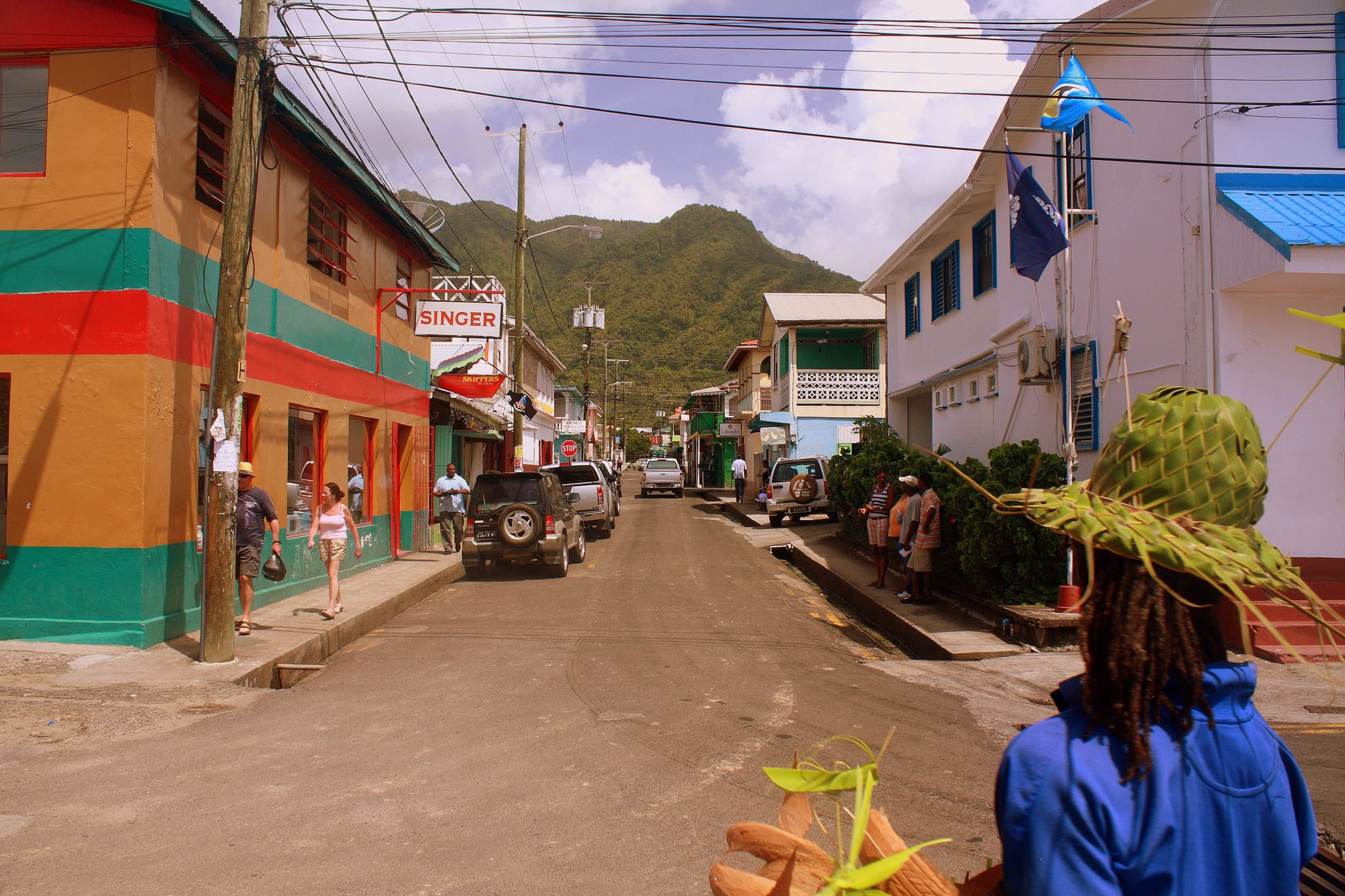 A colourful street in Soufrière lined with painted wooden shopfronts and homes, people walking past parked cars, forested mountains behind