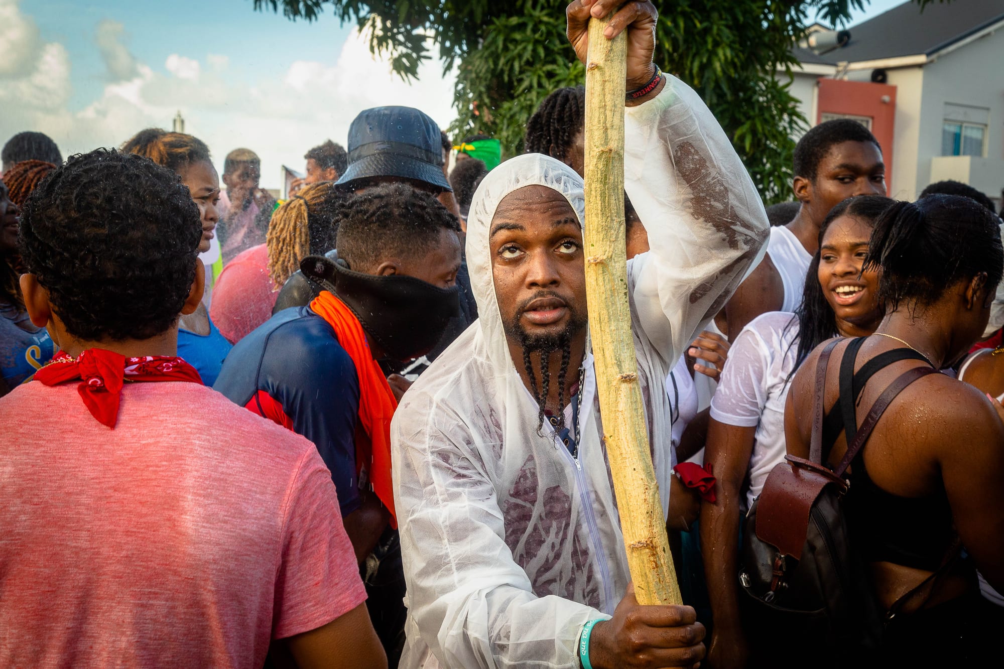 A crowd of young revellers in a street, one man in a white hooded plastic poncho holding up a long wooden stick, others wearing red scarves and wet T-shirts