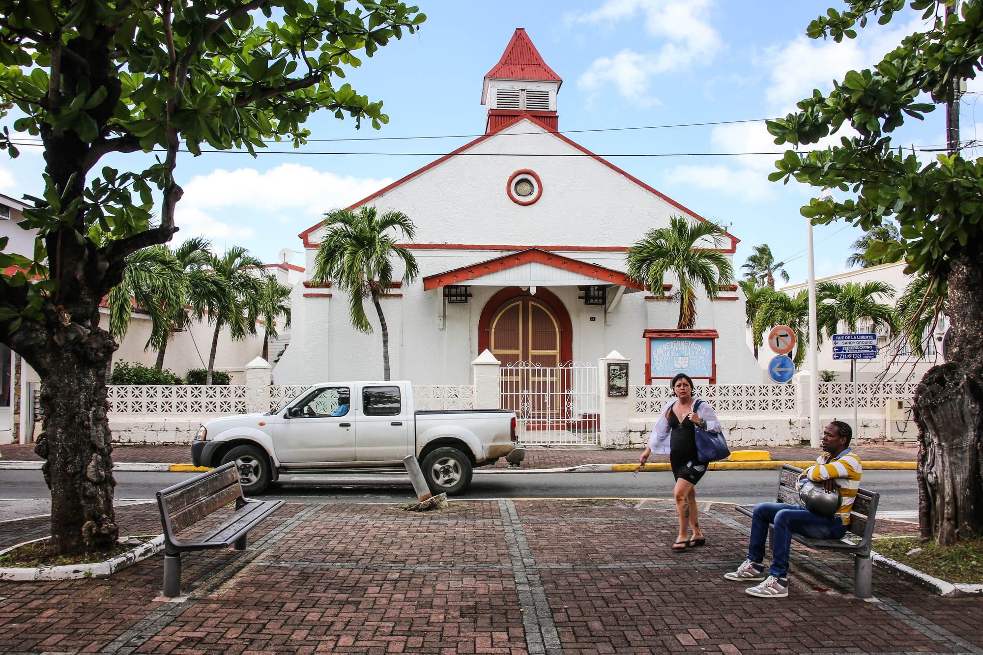 A white church with red trim, an arched wooden door under a red porch roof and a small red-roofed bell turret, set behind a white patterned wall with palm trees, as a woman walks past and a man sits on a bench