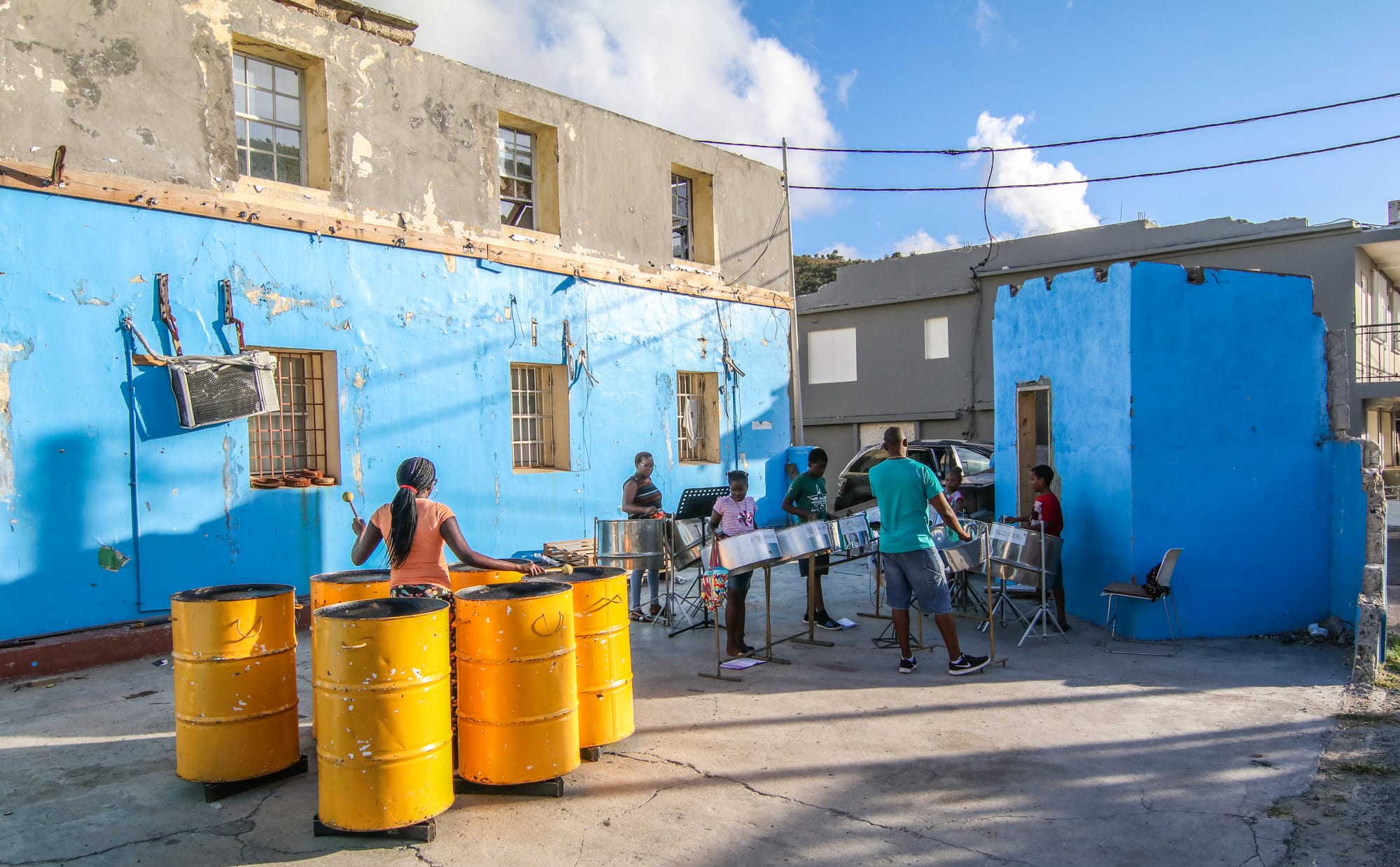 Young musicians practising on steel pans and yellow oil-drum bass pans in an open courtyard in front of a bright blue building with a damaged upper storey