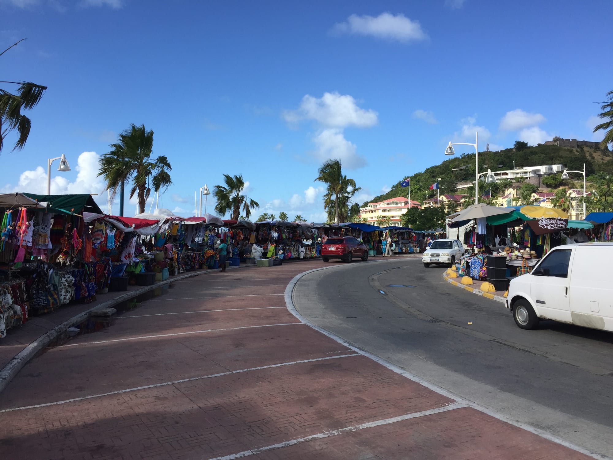 Rows of open-air market stalls with colourful awnings lining a curved waterfront road in Marigot, with a hilltop fort visible in the distance