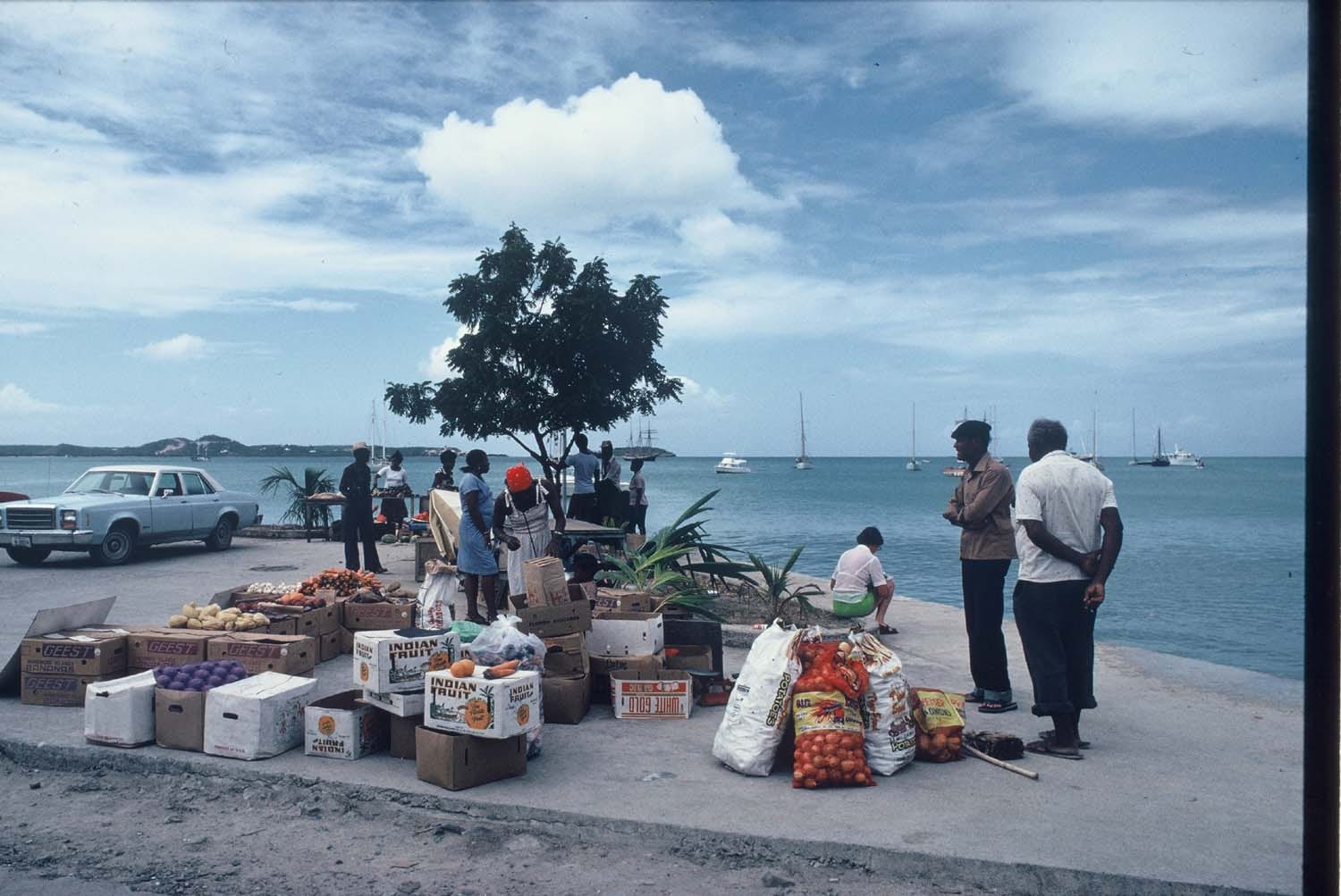 Boxes and sacks of fruit and vegetables laid out for sale on a quayside under a small tree, with shoppers, an old saloon car and yachts anchored in the bay behind