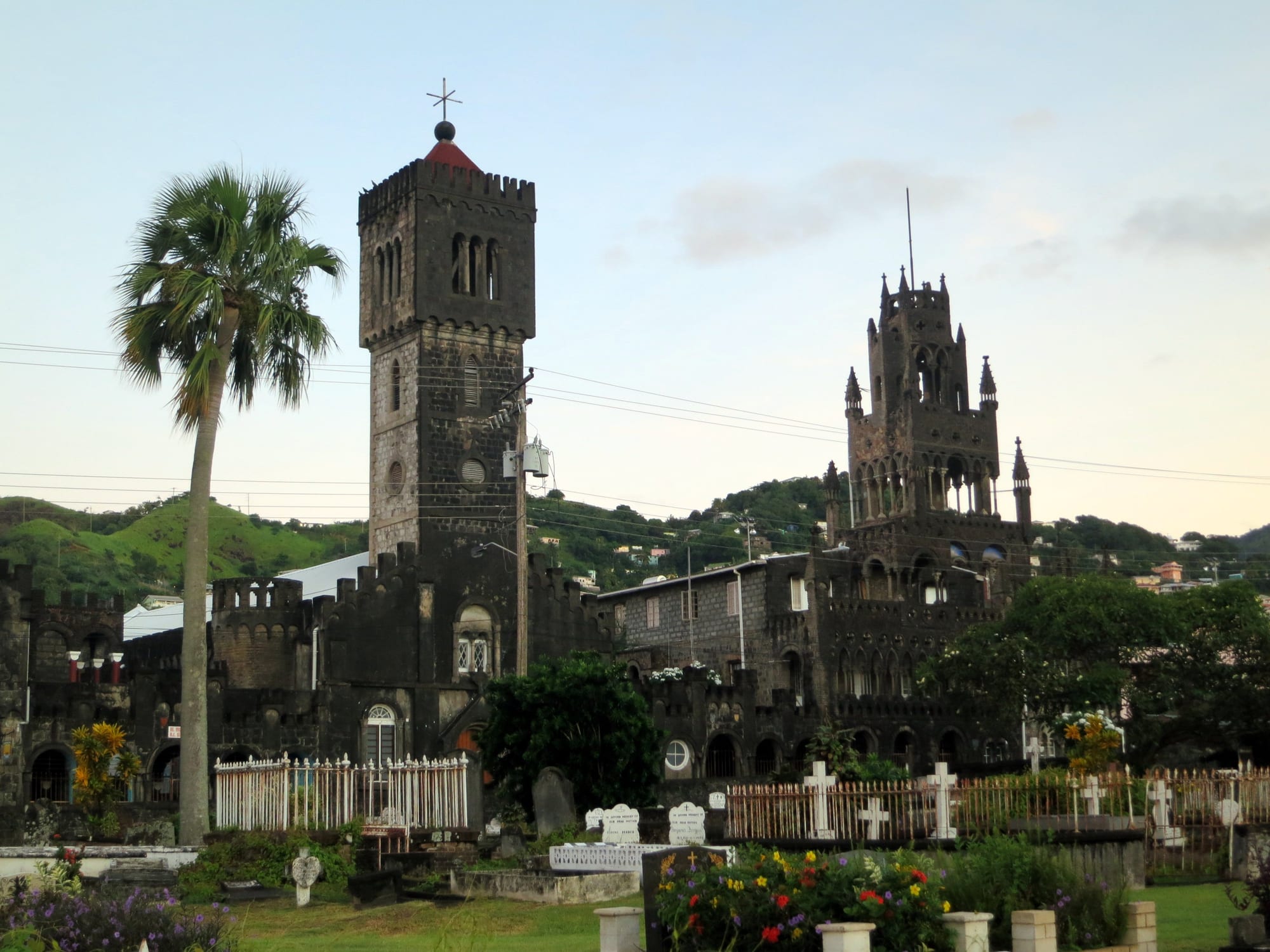 The dark stone bell tower and ornate pinnacled tower of a Catholic cathedral rising above a churchyard of white crosses and flowers, with a palm tree and green hills behind