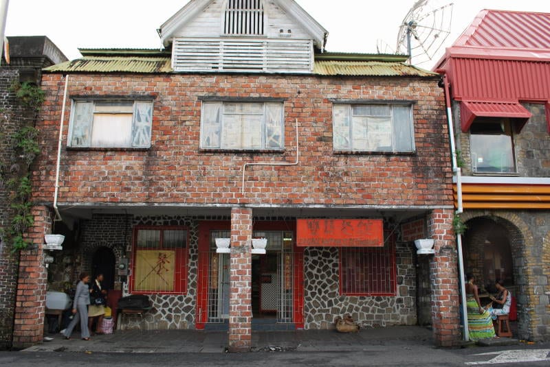 A weathered brick shopfront building in Kingstown with a Chinese-run store and pedestrians walking past on the street