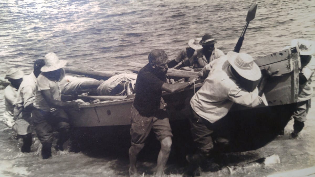 A black-and-white photograph of men in sun hats hauling a wooden open whaleboat up onto a beach through the surf