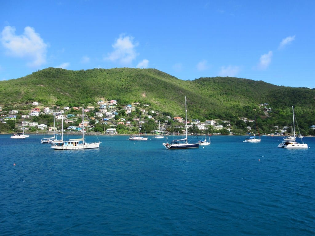 Sailing yachts and a catamaran anchored in a calm blue bay below a green hillside dotted with houses