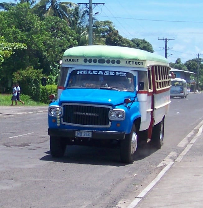 A brightly painted blue, white and cream wooden bus, decorated with the words RELEASE ME, driving along a road lined with palm trees