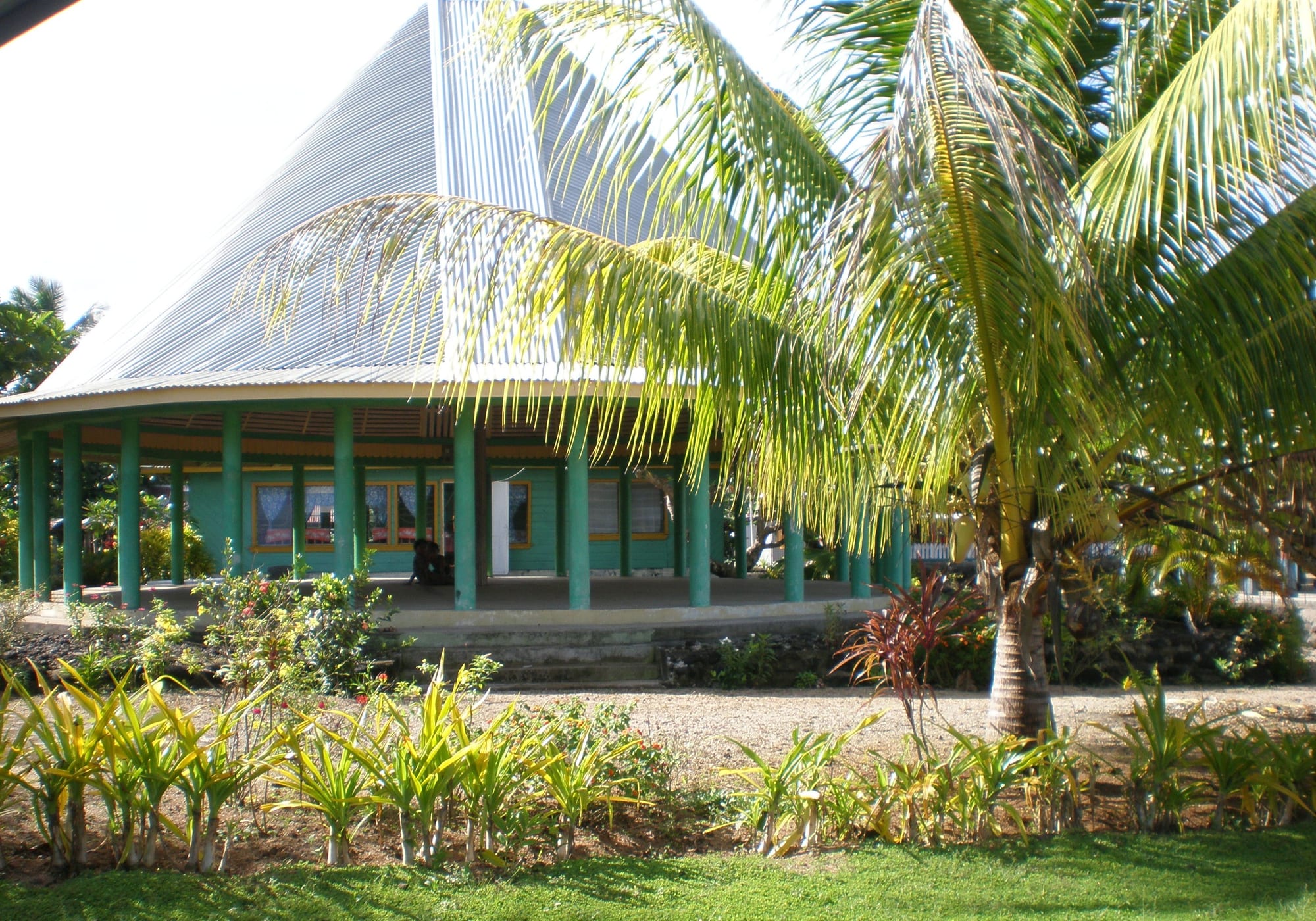 A round, open-sided traditional Samoan house with green pillars and a peaked corrugated roof, set among palm trees and gardens