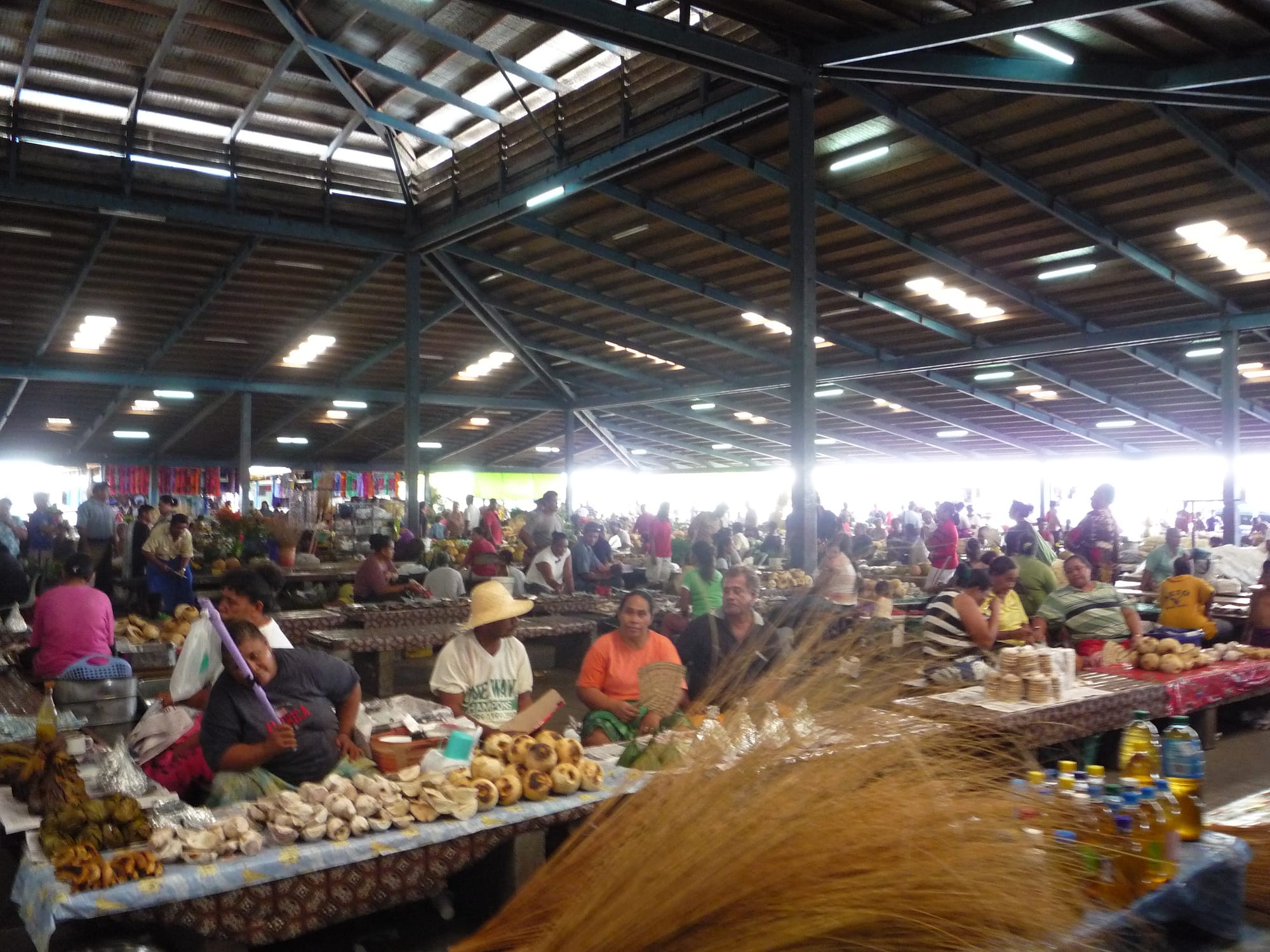 A crowded covered market hall in Apia with sellers sitting behind tables of coconuts, bananas and woven goods under a high steel roof