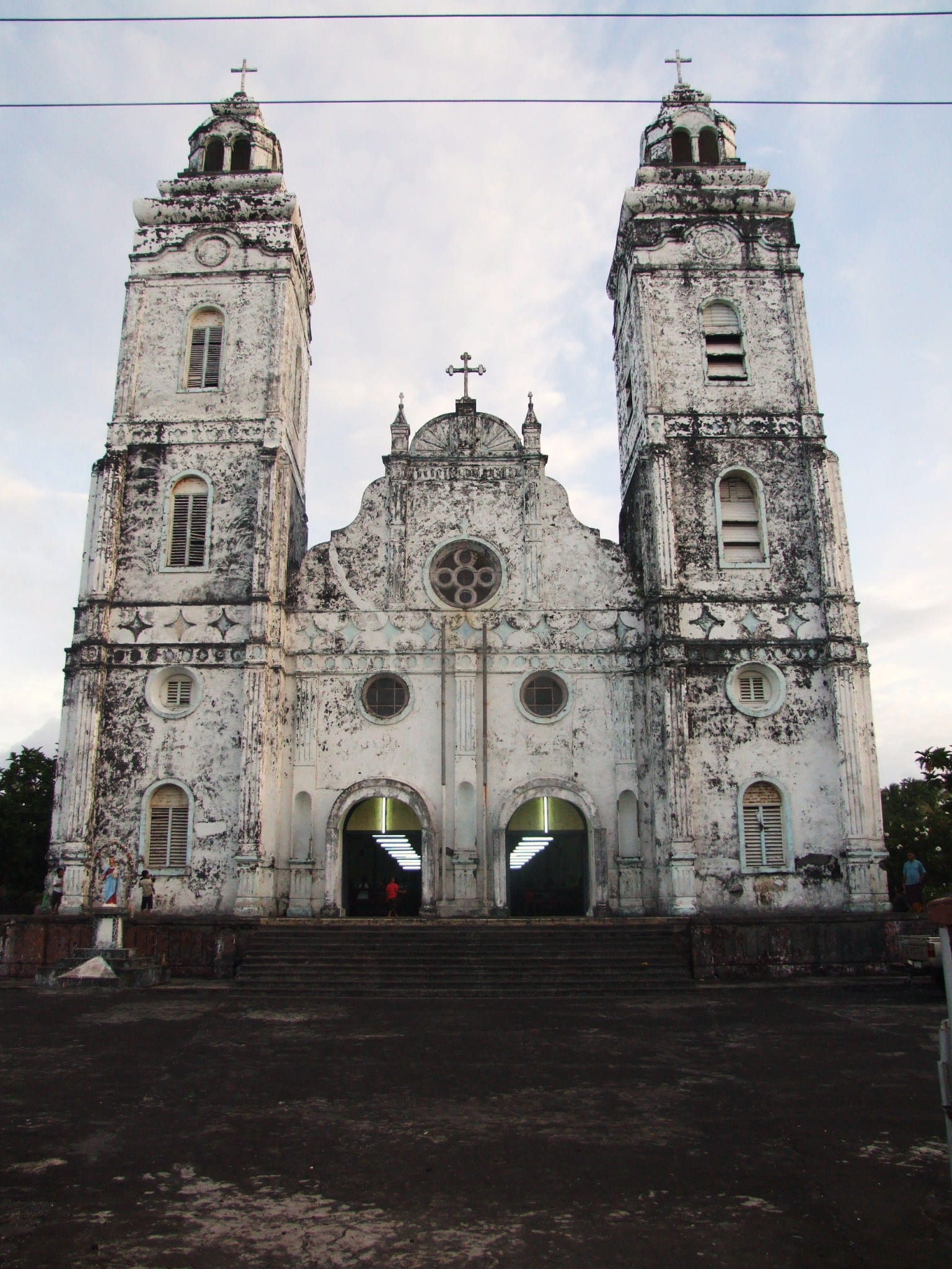 The weathered white facade of a large twin-towered church with arched doorways lit from within, crosses on each tower and a wide open forecourt in front