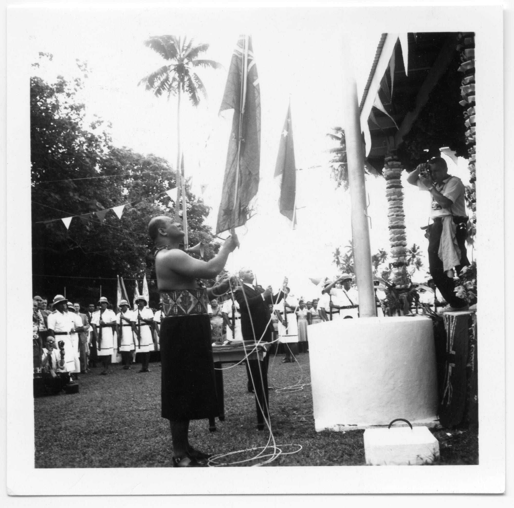 A black and white photograph of a Samoan leader in a patterned wrap and a man in a dark suit lowering flags from a flagpole, with uniformed men in white standing in lines behind and a photographer on a raised platform