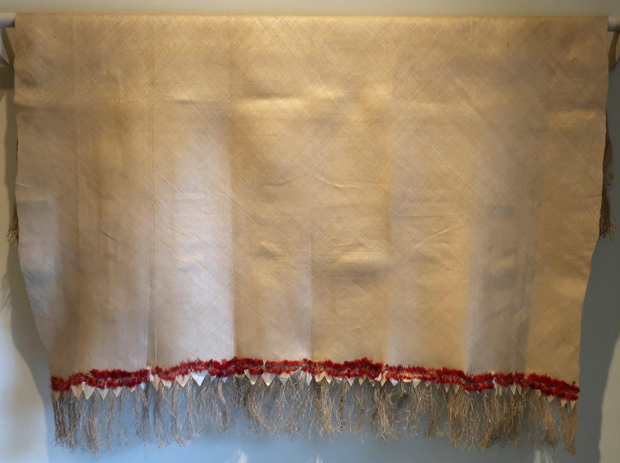 A large, pale, finely woven pandanus mat hanging from a rod against a white wall, with a band of red feathers along the lower edge above a long loose fringe