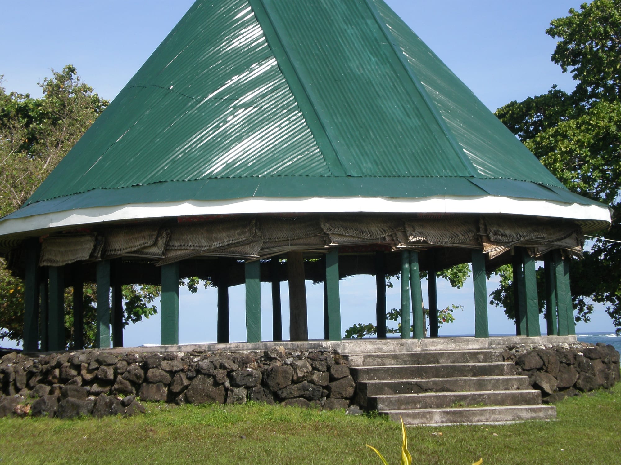 A round open-sided house with a tall conical green roof on wooden posts, with woven blinds rolled up under the eaves, standing on a raised stone platform with steps, the sea visible between the posts