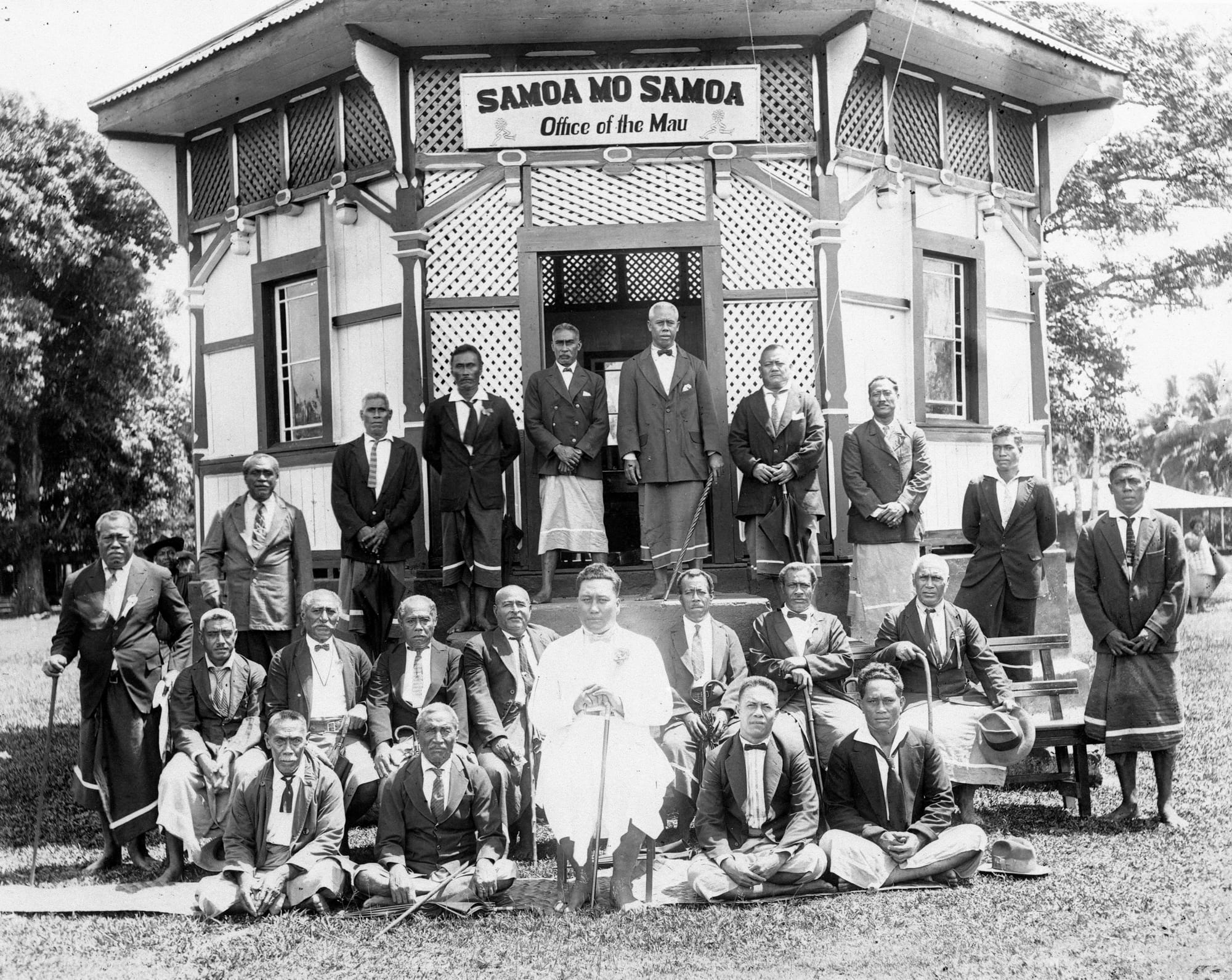 A group of Samoan Mau movement leaders in formal dress and lavalava seated and standing outside a building in 1929