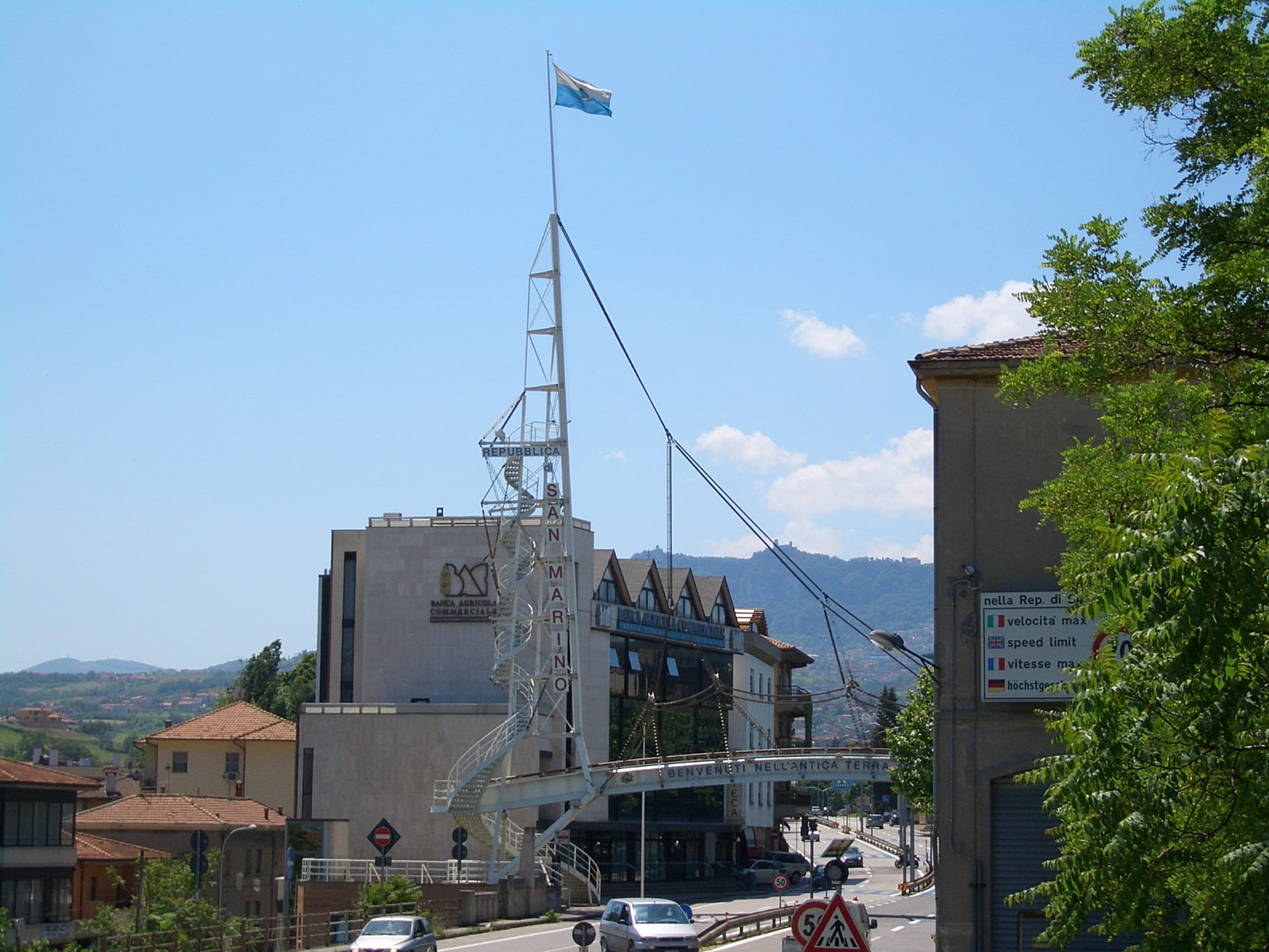 A road arch reading Benvenuti nell'antica terra della liberta, spanning a street lined with bank buildings, with San Marino's hilltop city visible on a hill behind