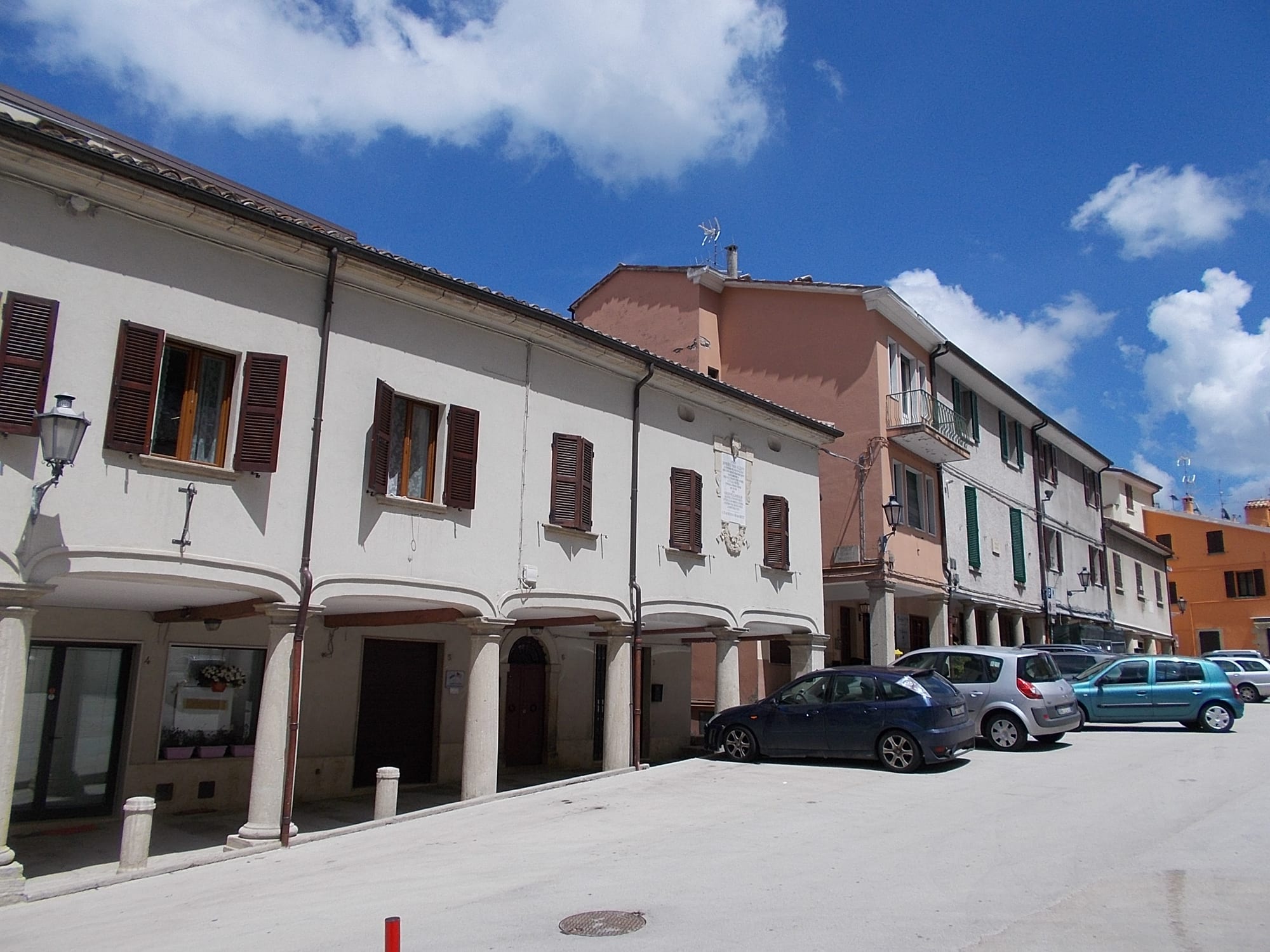 An ordinary residential street of shuttered stone houses with parked cars, under a blue sky with clouds