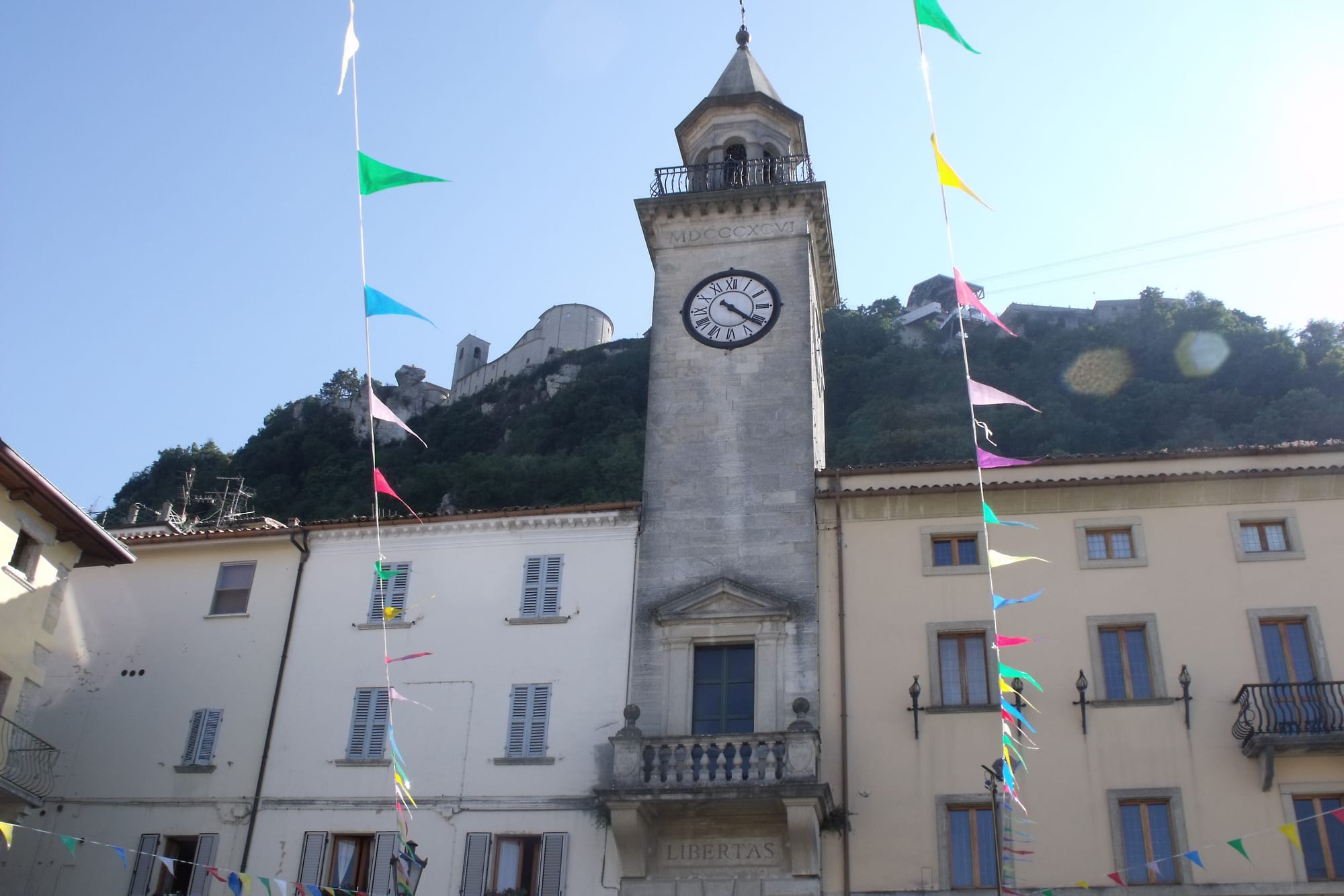 A stone clock tower marked LIBERTAS in a town square, decorated with coloured festival flags, with San Marino's fortress-topped hill rising behind it