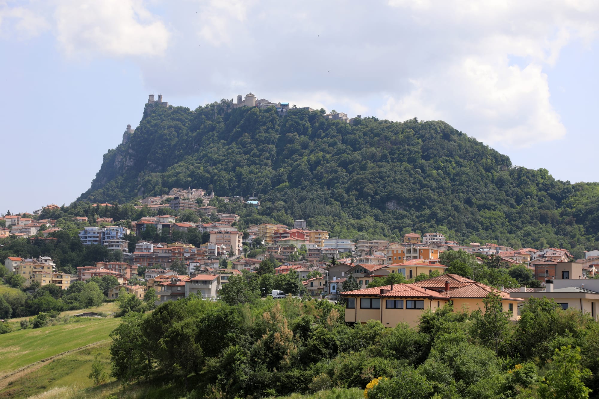 A steep wooded mountain with fortifications along its crest, rising above a spread of red-roofed houses and green fields