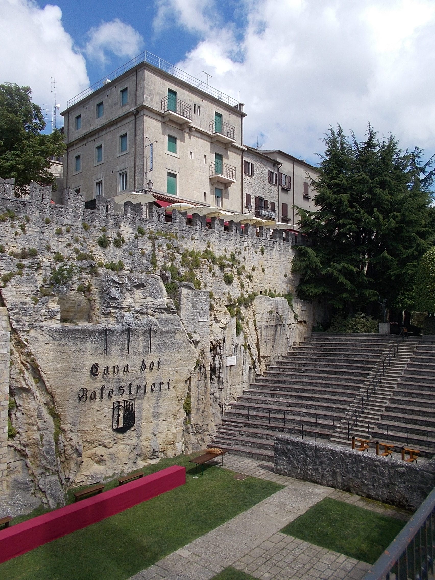 A grassy arena at the foot of a high stone wall and cut rock face carved with the words 'Cava dei Balestrieri' and a coat of arms, with tiers of stone steps rising to the right and buildings above