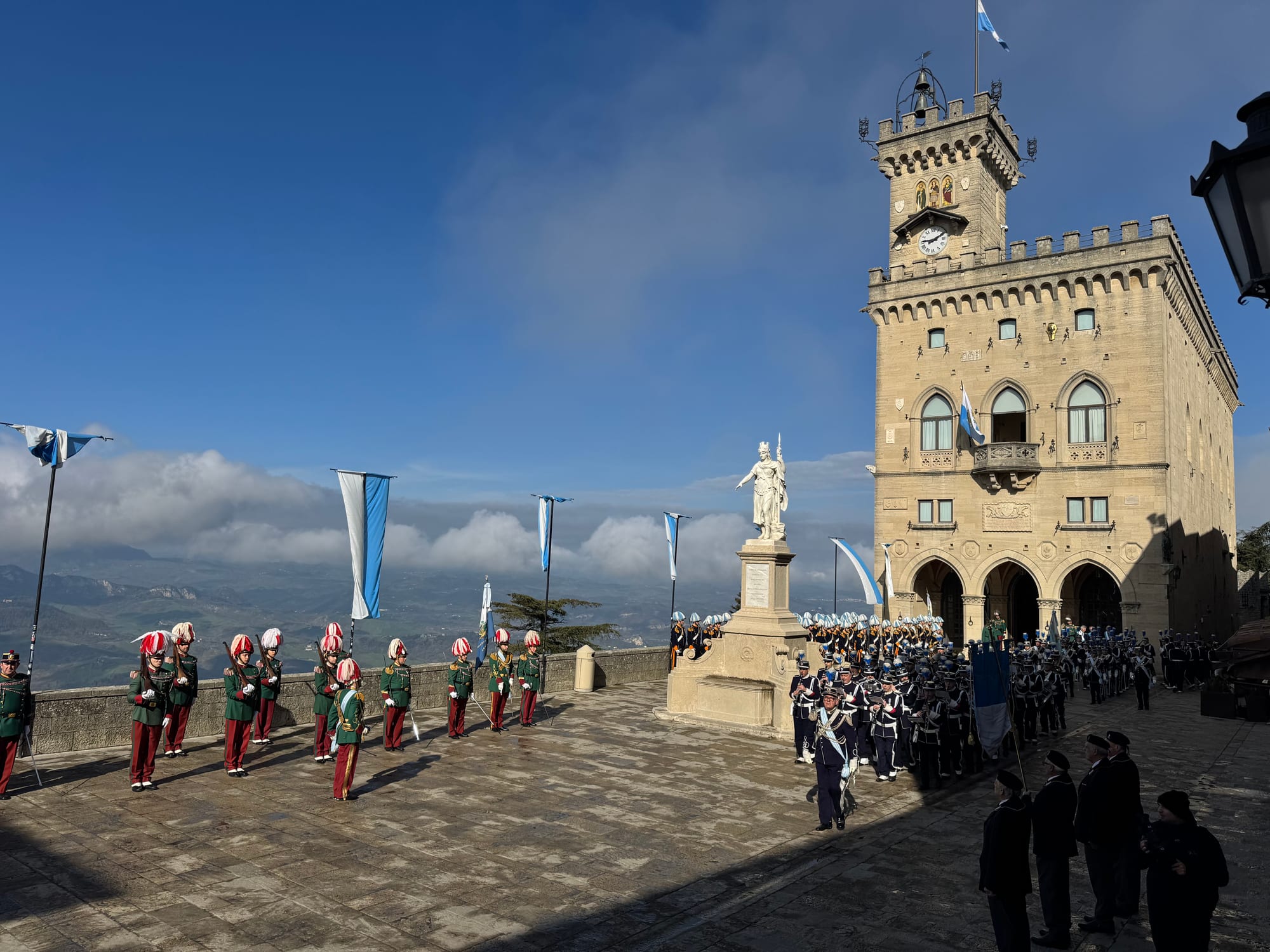 Ranks of uniformed guards and musicians in plumed helmets parade on a stone square beside a white statue, in front of a crenellated palace with a clock tower, with blue and white flags and clouds over the valley beyond