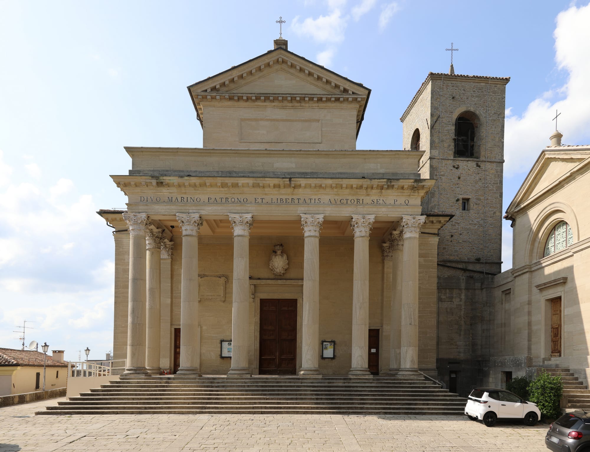 A pale stone neoclassical church with a portico of tall Corinthian columns above a wide flight of steps, a Latin inscription along the frieze and a square stone bell tower to the right