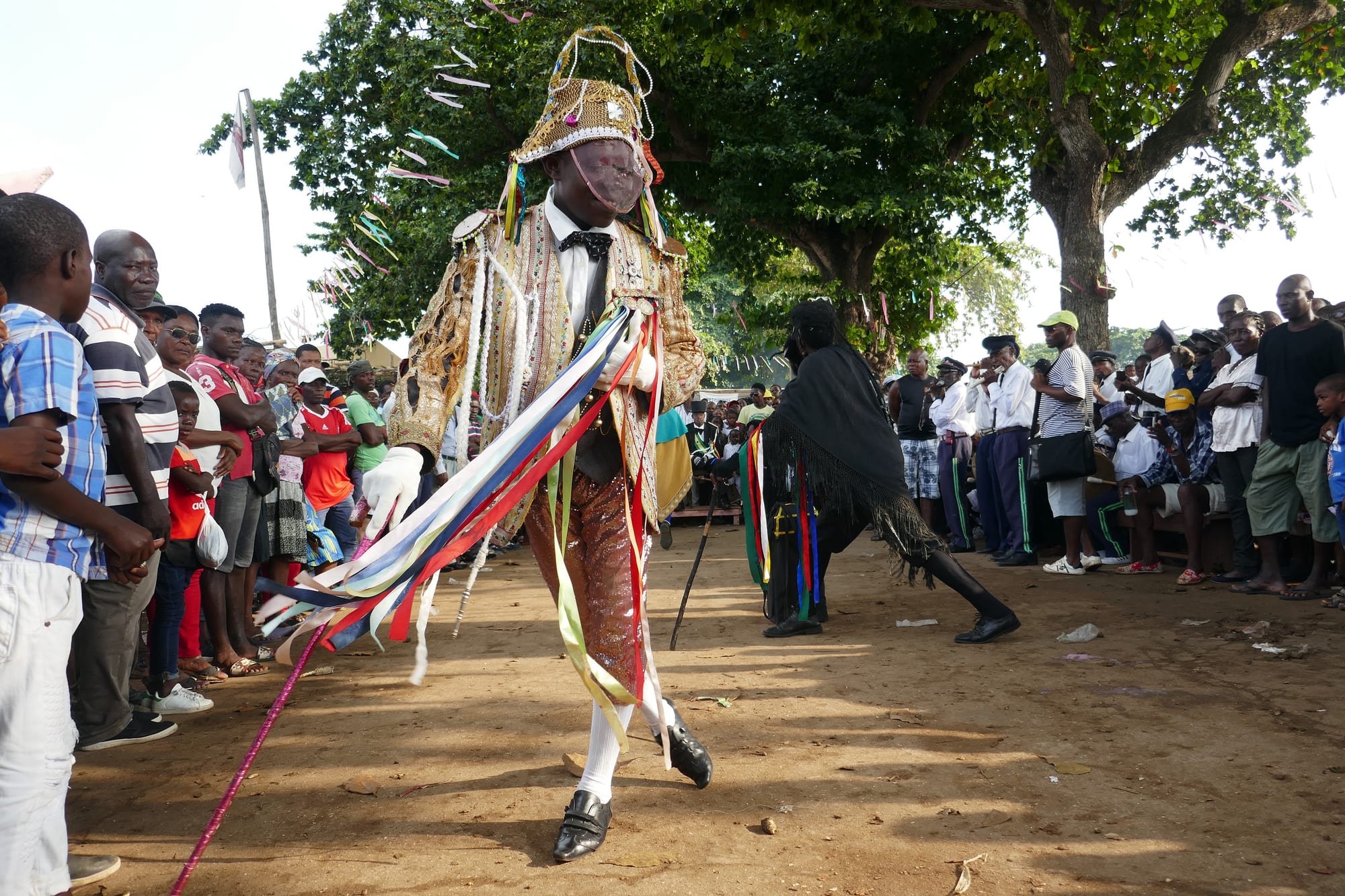 A tchiloli performer in a gold embroidered coat, pale mesh mask and plumed hat trails long coloured ribbons as he dances across a dirt square, watched by a crowd and a row of drummers under the trees