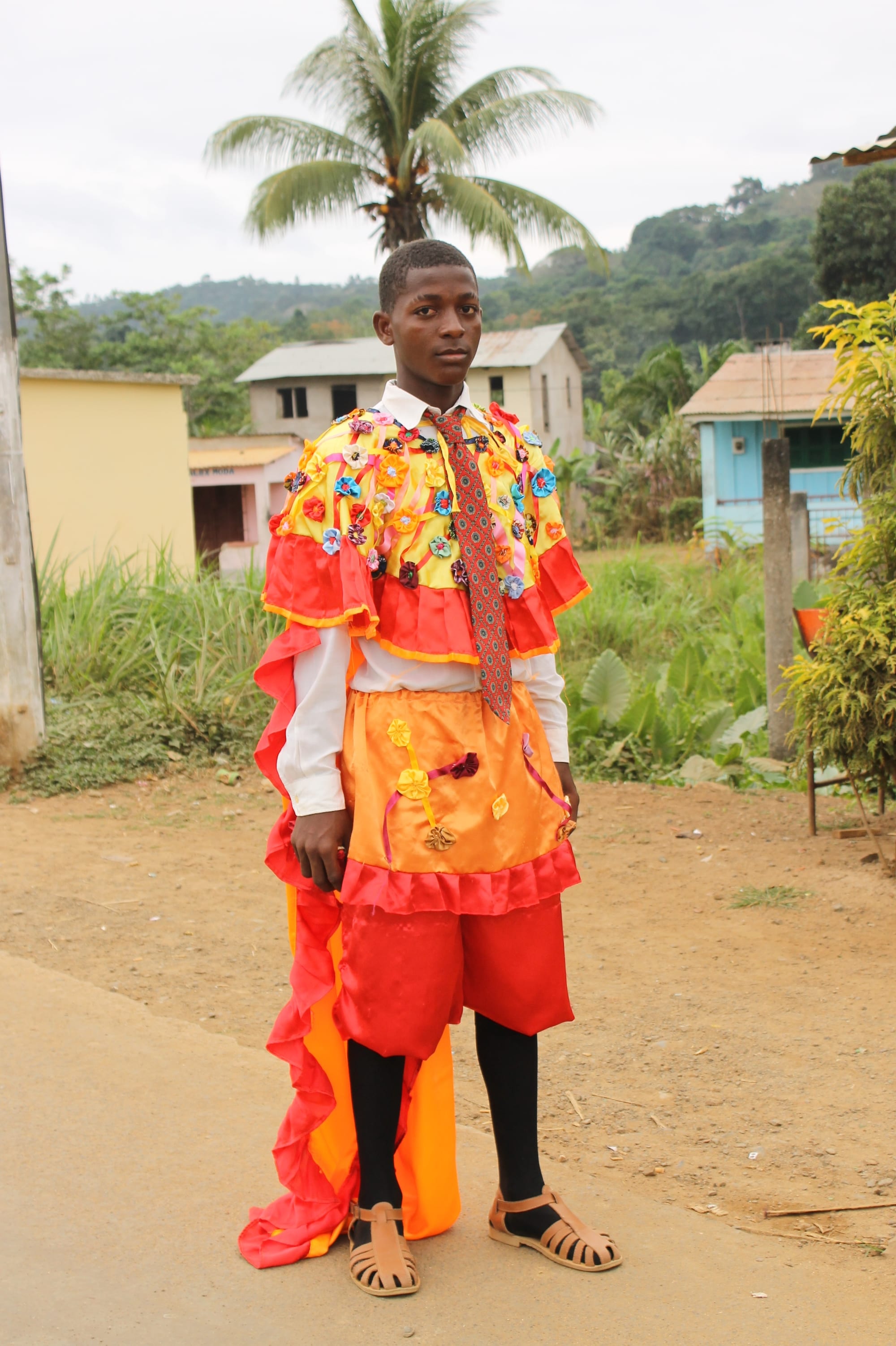 A young man stands on a dirt road in a costume of red and orange satin with ruffled edges and a long ruffled train, a yellow cape covered in coloured fabric rosettes, a white shirt and patterned tie, black stockings and sandals, with houses, a palm tree and green hills behind