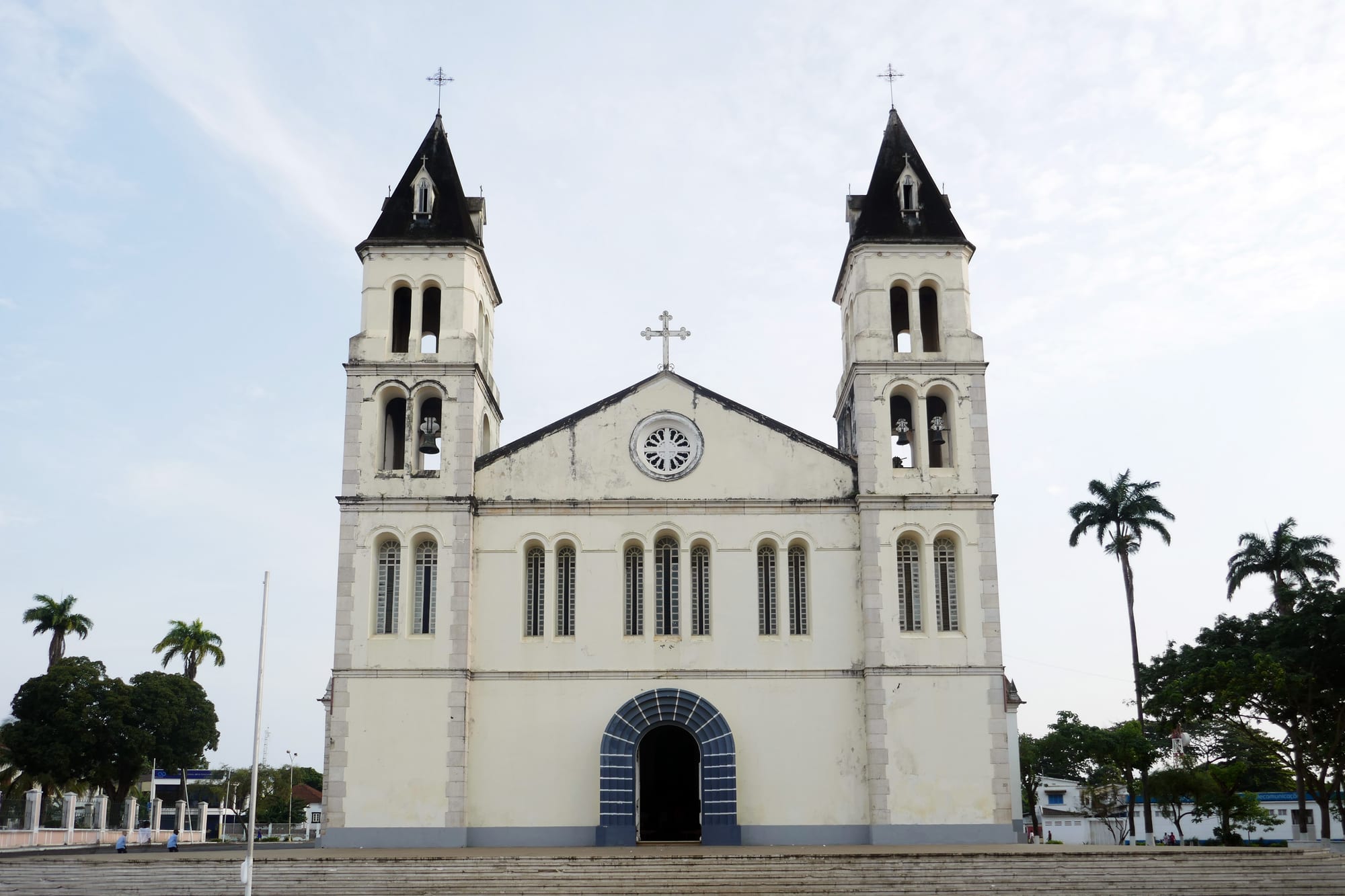 The pale cream facade of a cathedral with two bell towers topped by dark pointed spires, a rose window and a blue arched doorway, beneath a pale sky with palm trees at the sides