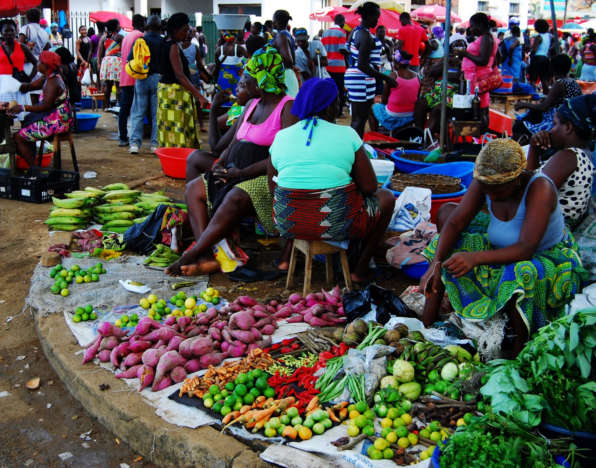 A crowded outdoor market with women selling limes, sweet potatoes, carrots and plantains spread on cloths on the ground