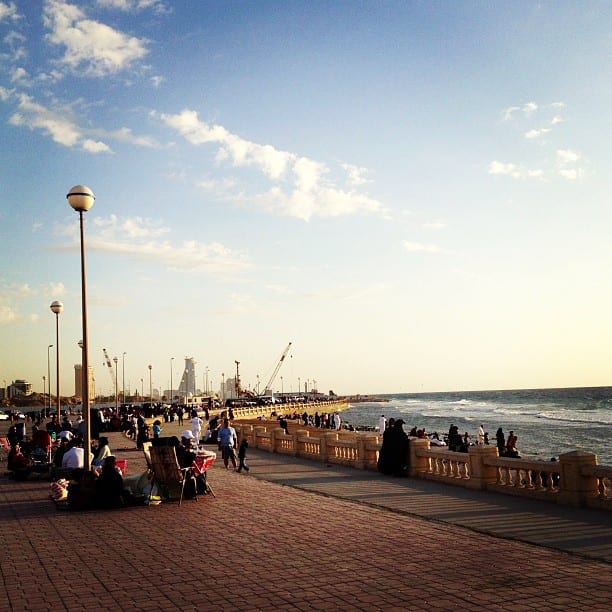 A crowd of people sitting and walking along a seaside promenade at sunset, with a stone balustrade and the sea beyond