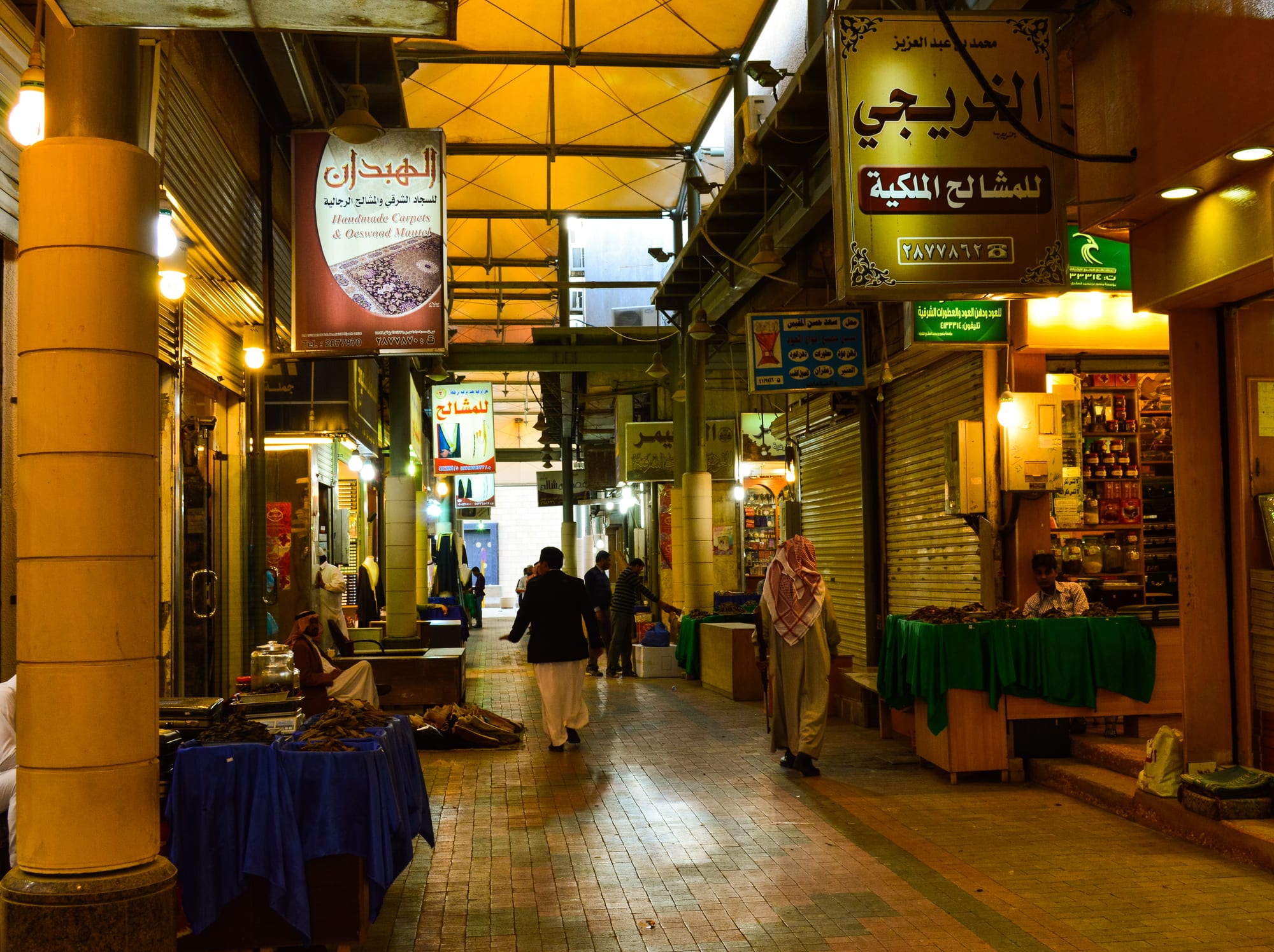 A covered market alley in downtown Riyadh at dusk, lit by shop signs and hanging lamps, with vendors and shoppers among stalls of goods