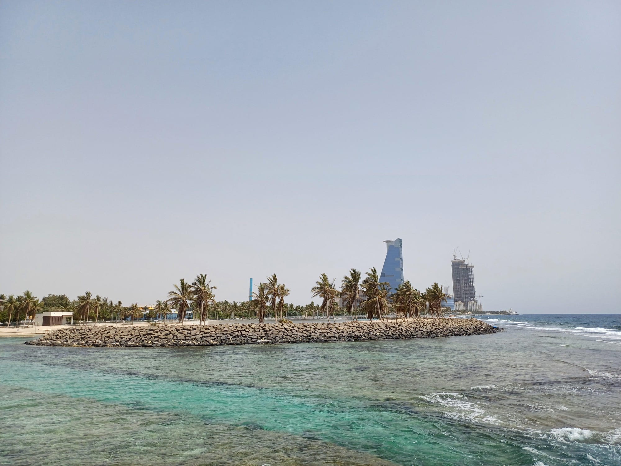 Turquoise Red Sea water in front of a palm-lined rock breakwater and city towers
