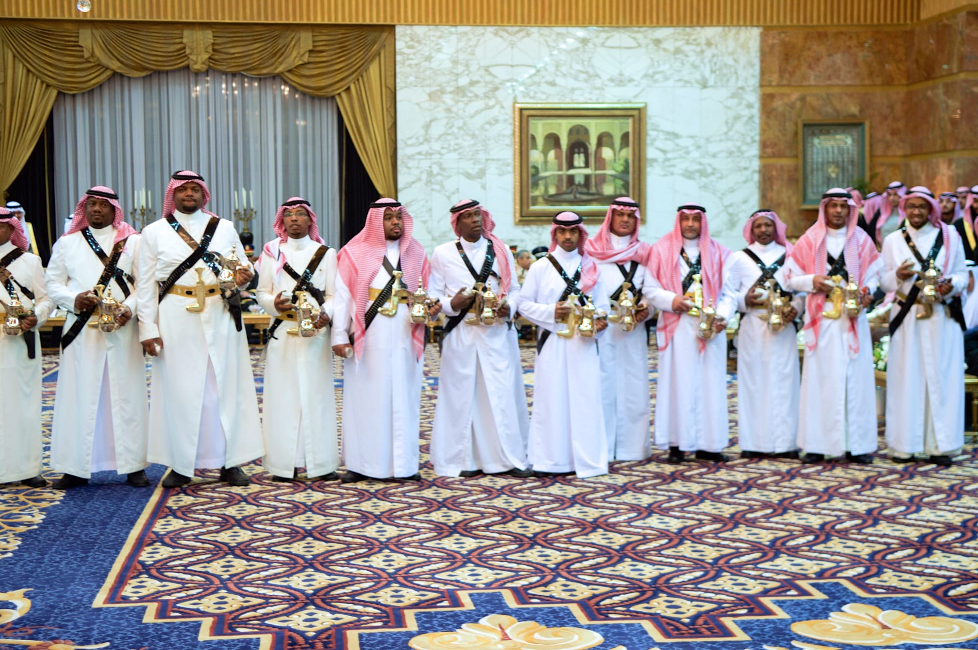 A line of men in white thobes and red-checked headscarves, each wearing a crossed leather sash and holding brass coffee pots and small cups, stand on a patterned carpet in a large reception hall